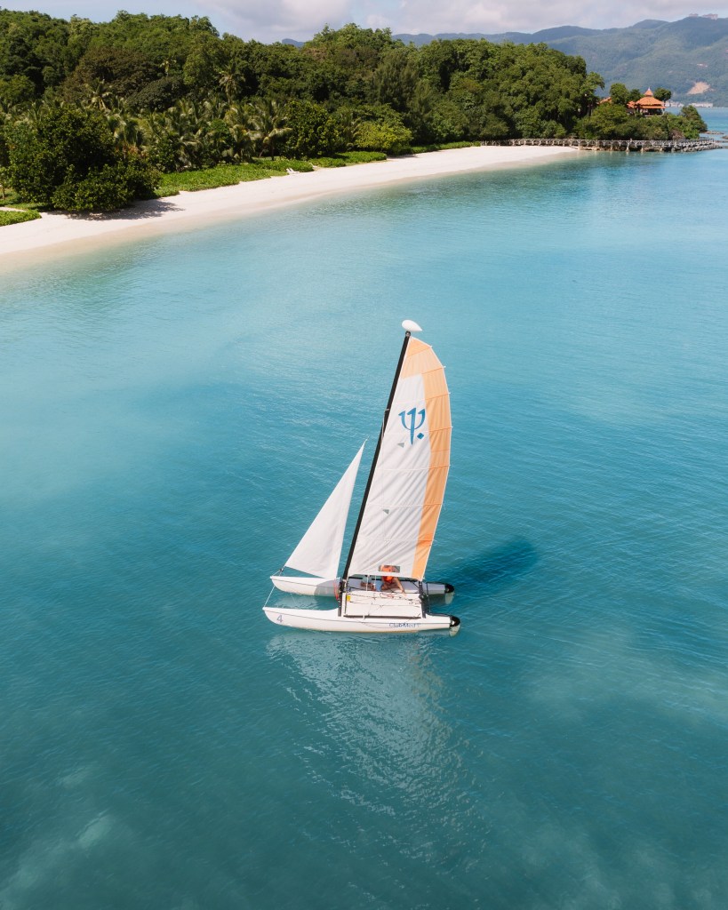 A catamaran sailboat with orange and white sails sailing on turquoise waters near a sandy beach and lush greenery.