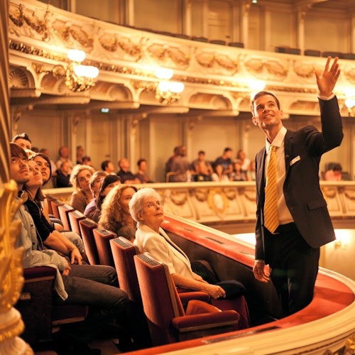 Opas elehtii puhuessaan istuvalle yleisölle koristeellisessa teatterissa, jossa on parvekkeita ja kattokruunuja.
