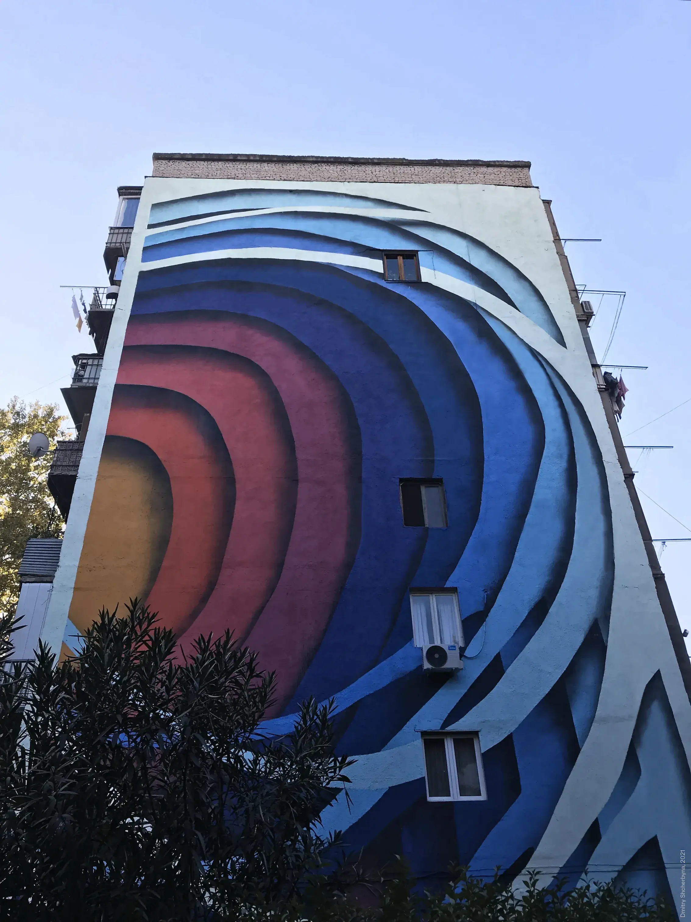 A mural covering the side of an apartment building features bold concentric curved bands that shift from warm gold and red in the center to deep blue on the outer edges, creating an optical illusion of depth.