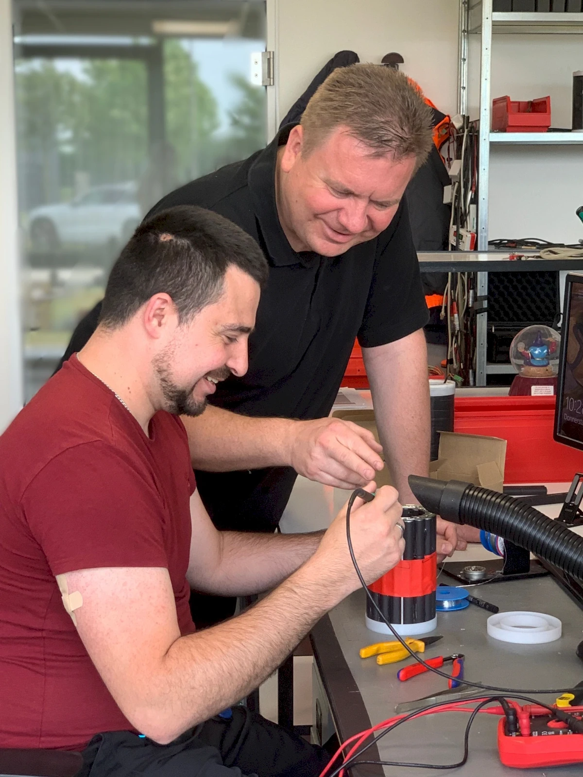 Two men collaborate on electrical device repair, with one holding a multimeter near a battery pack.