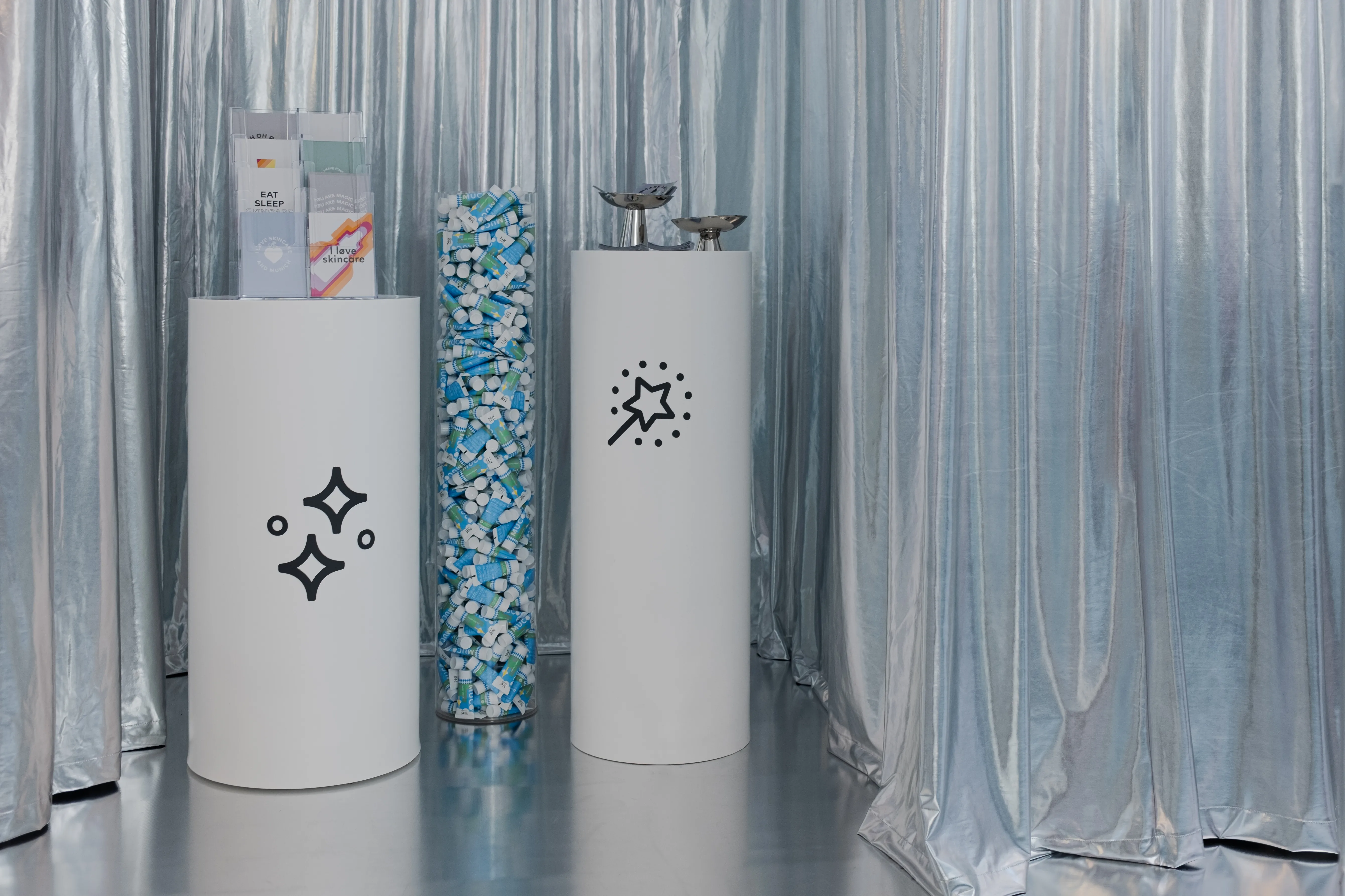 White pedestals with sparkle icons and a clear column filled with colorful capsules in front of silver curtains at the NØ Cosmetics pop-up.
