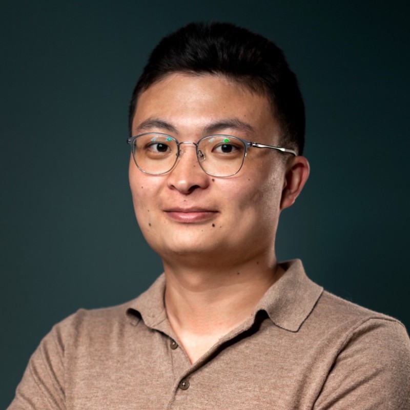 Portrait of a person with red hair, wearing a tan coat and white turtleneck, set against a gray background.