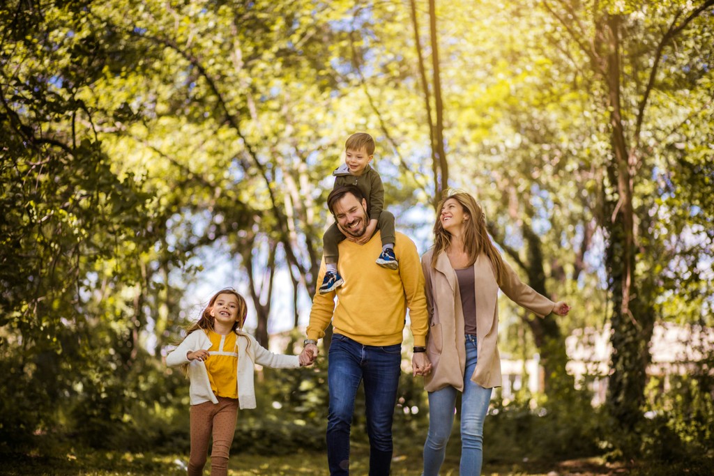 A happy family enjoying a sunny day in a lush forest, with a father carrying his young son on his shoulders, a mother walking alongside, and a daughter joyfully holding her father's hand.