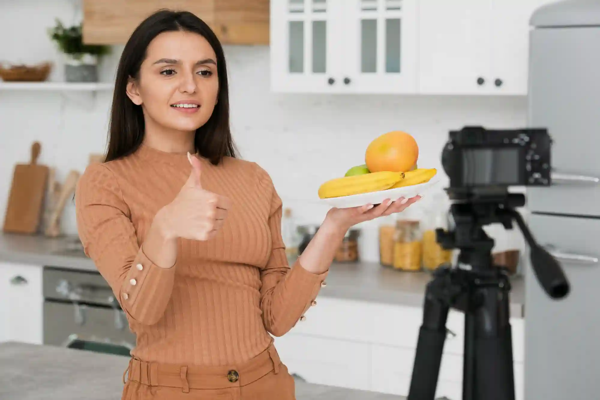 Woman in a kitchen recording a video, holding a plate of fruit and giving a thumbs-up gesture to the camera.