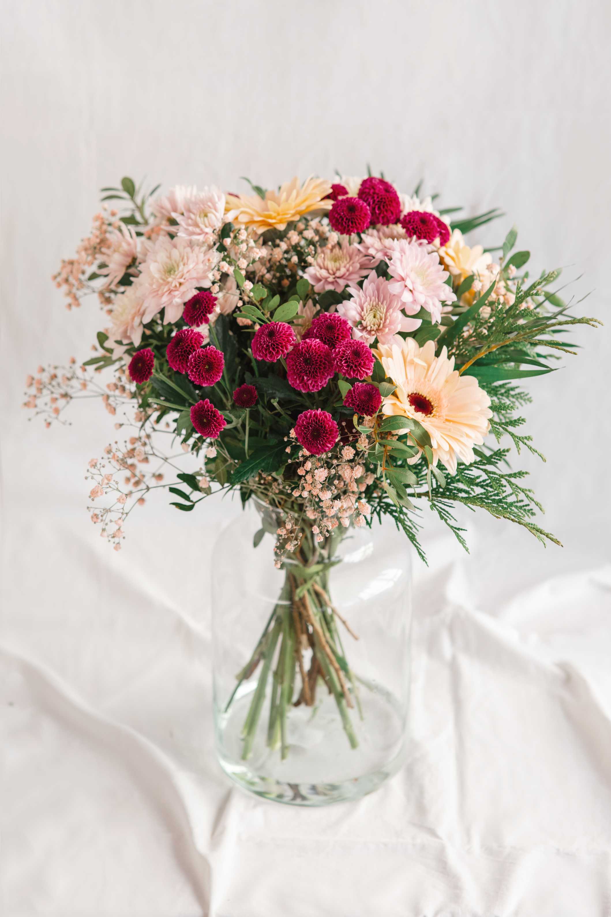 Ram de flors en gerro de vidre amb gerberes préssec, crisantems rosa i fúcsia, paniculata i verds decoratius, sobre fons clar.