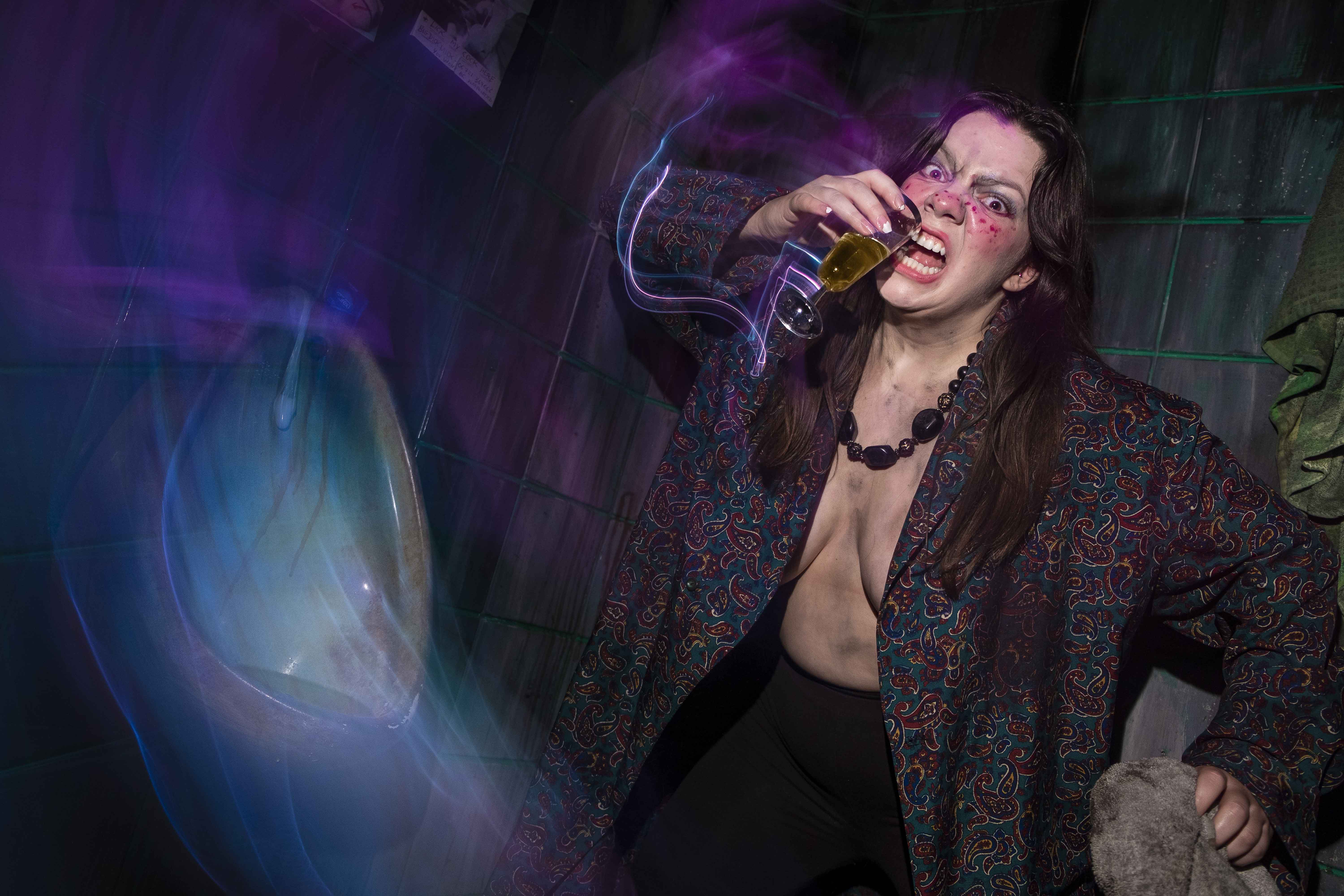 A person in a patterned jacket and bold makeup energetically poses with a drink in front of artistic swirling lights and a decorated wall, creating a dynamic and colorful scene.