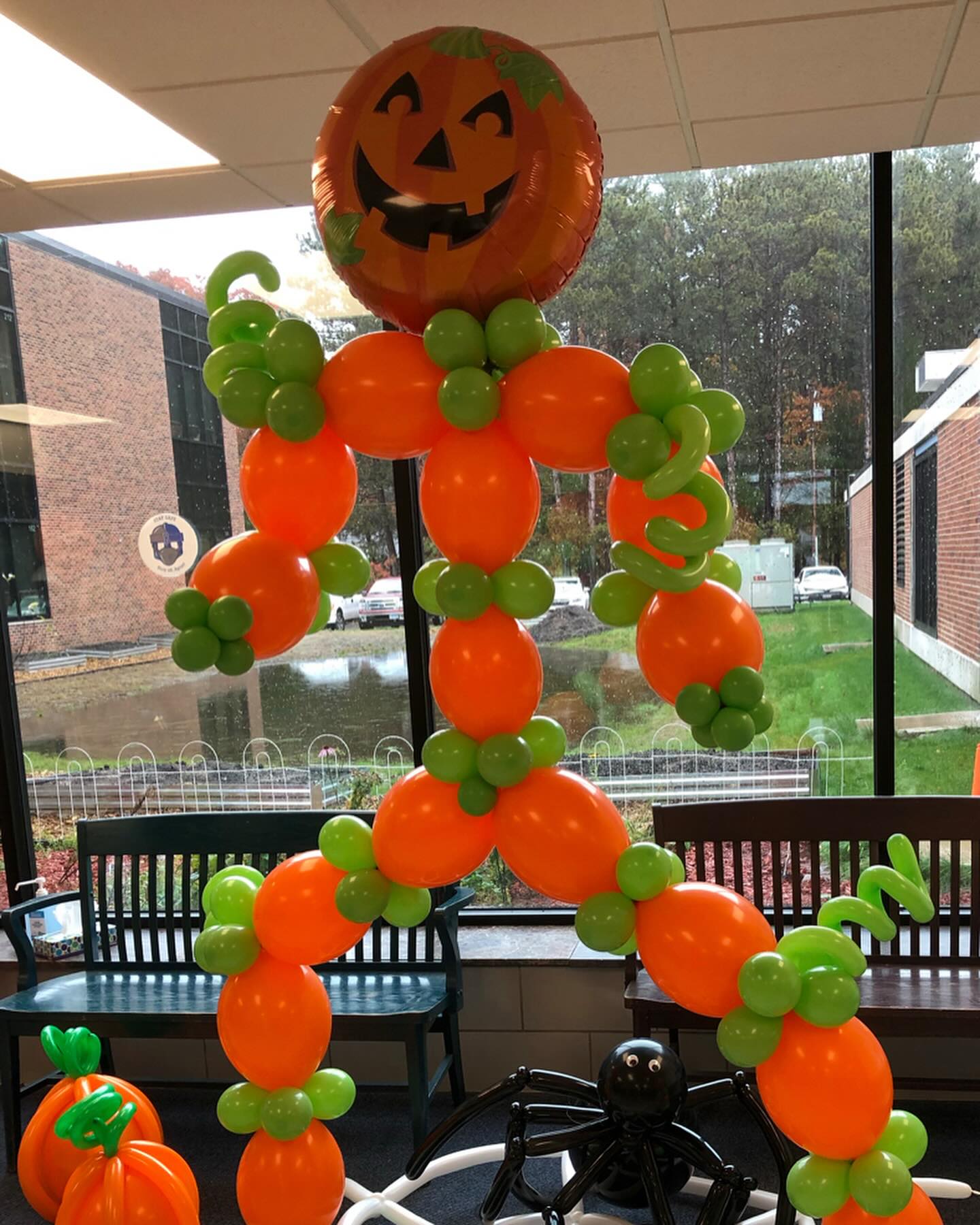 A large Halloween-themed balloon sculpture resembling a pumpkin-headed figure with orange and green balloons stands inside a room with large windows, overlooking a parking lot and trees.