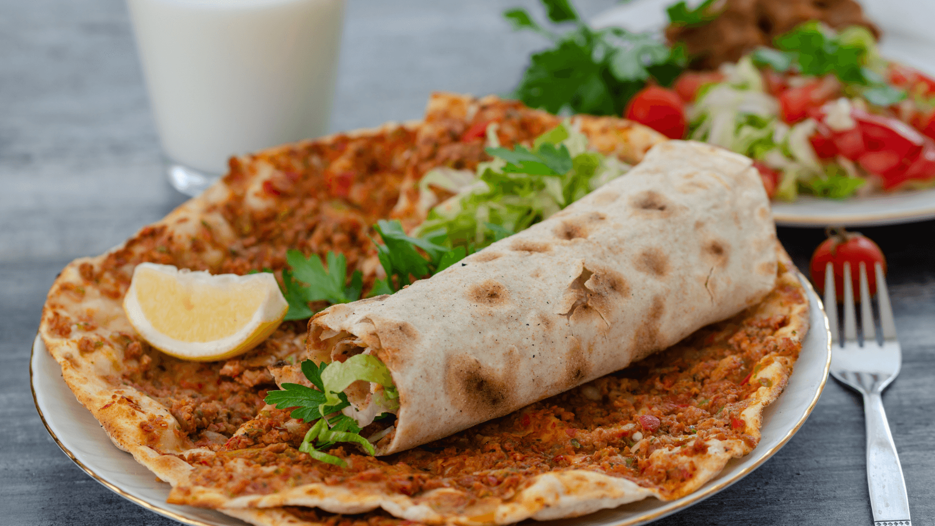 A traditional Turkish lahmacun served with fresh parsley, lettuce, and a slice of lemon, partially wrapped as a dürüm, accompanied by a glass of ayran and a side salad in the background.