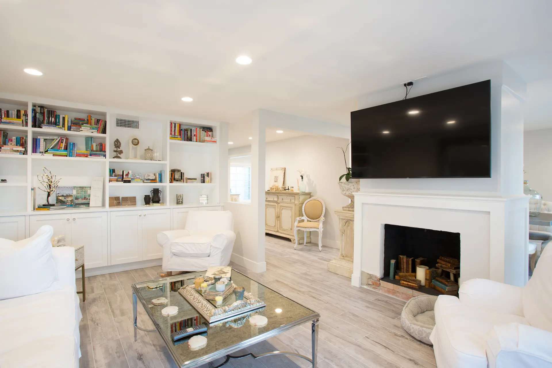 Elegant living room with gray hadwood floors, fireplace, and wall-mounted TV. Elegant dining area with large window, grey stone table, tufted chairs. From Dana Point Cape Cod remodel, photo by Chris Darnall