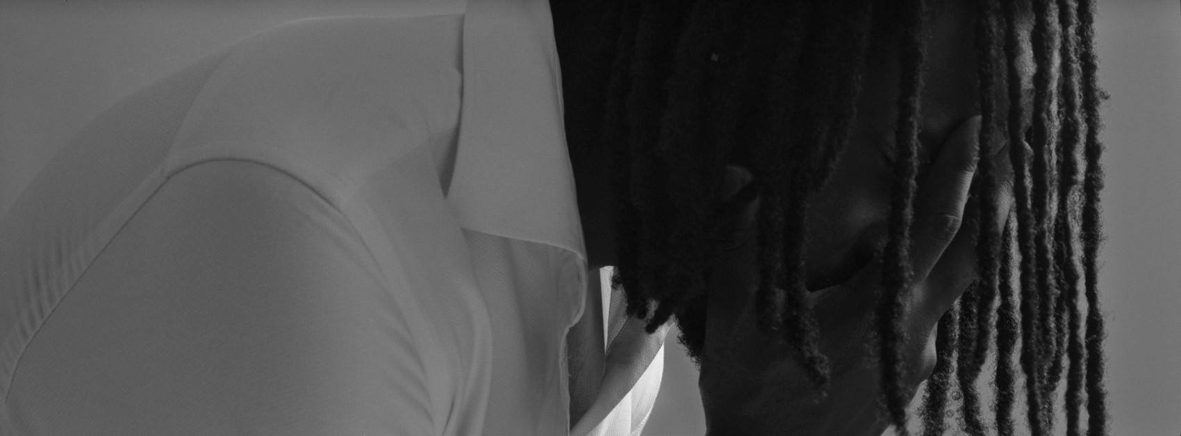 Film photography with flash: A dark skinned man sitting in a deep thinking position with his hair locks falling in front of his face. using Hasselblad XPan, wearing a white sweater with locks, close-up panoramic portrait of model in thought, black and white Ilford Delta. 