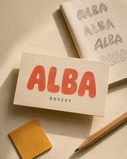 Business card with "ALBA BAKERY" in bold red and green text, pencil, sticky note, and notebook with "ALBA" logo sketches on a beige surface.