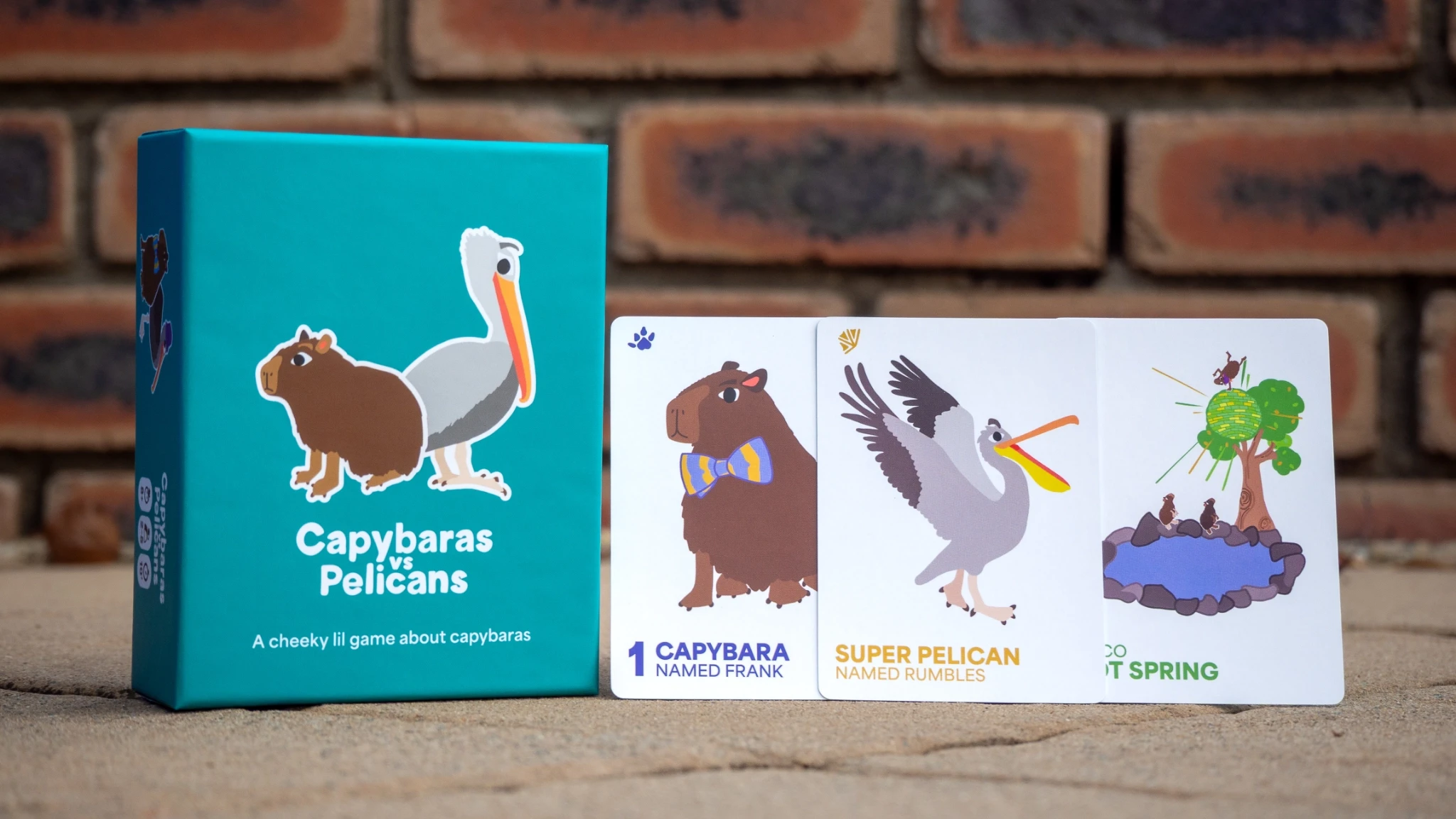Capybaras vs Pelicans board game box alongside three playing cards.