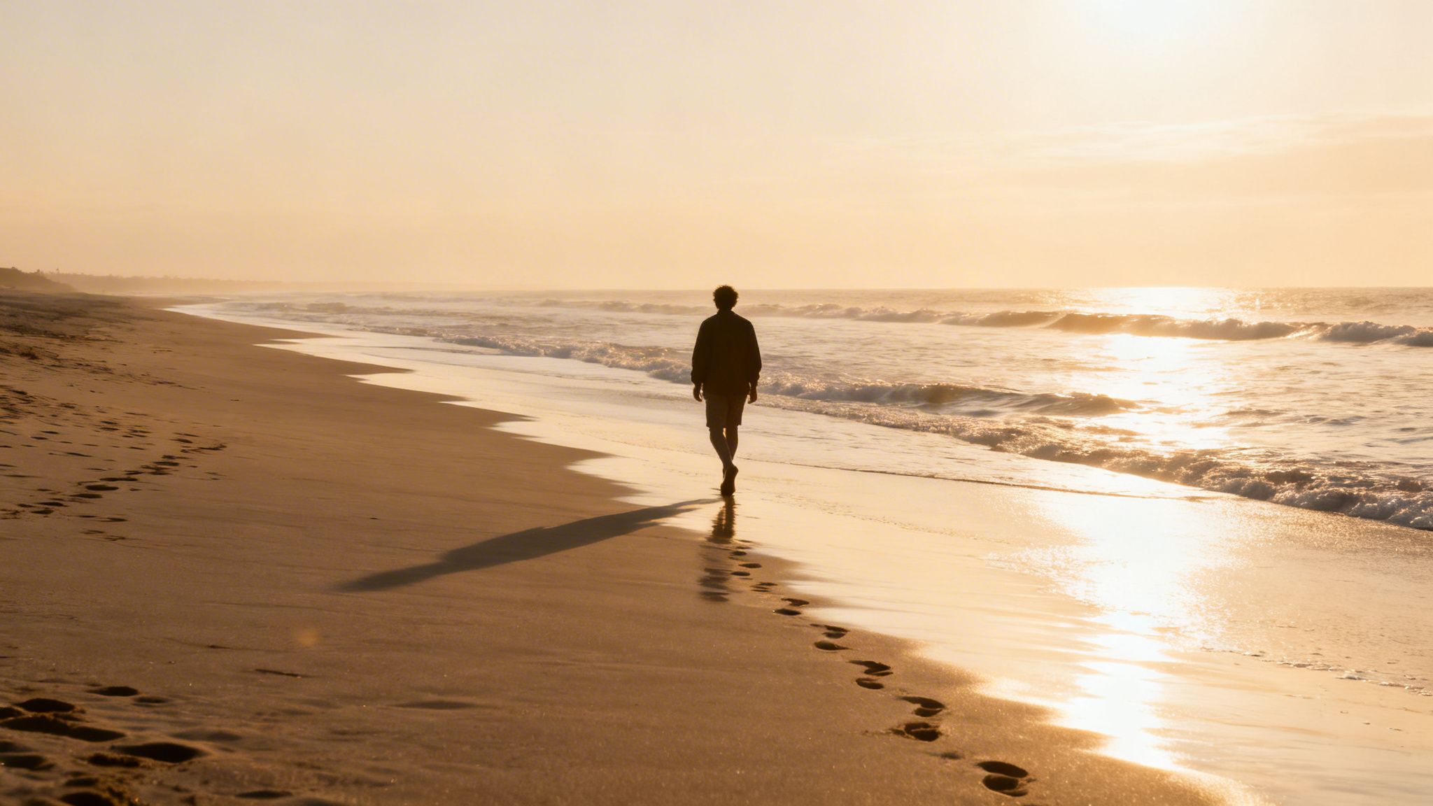 A lone person walks on a golden beach at sunset, leaving footprints in the wet sand.