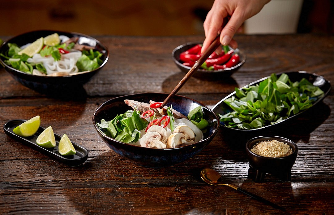 A hand uses chopsticks to prepare a bowl of fresh vegetable and noodle soup, surrounded by ingredients like lime wedges, red chili peppers, leafy greens, and a bowl of sesame seeds on a rustic wooden table.