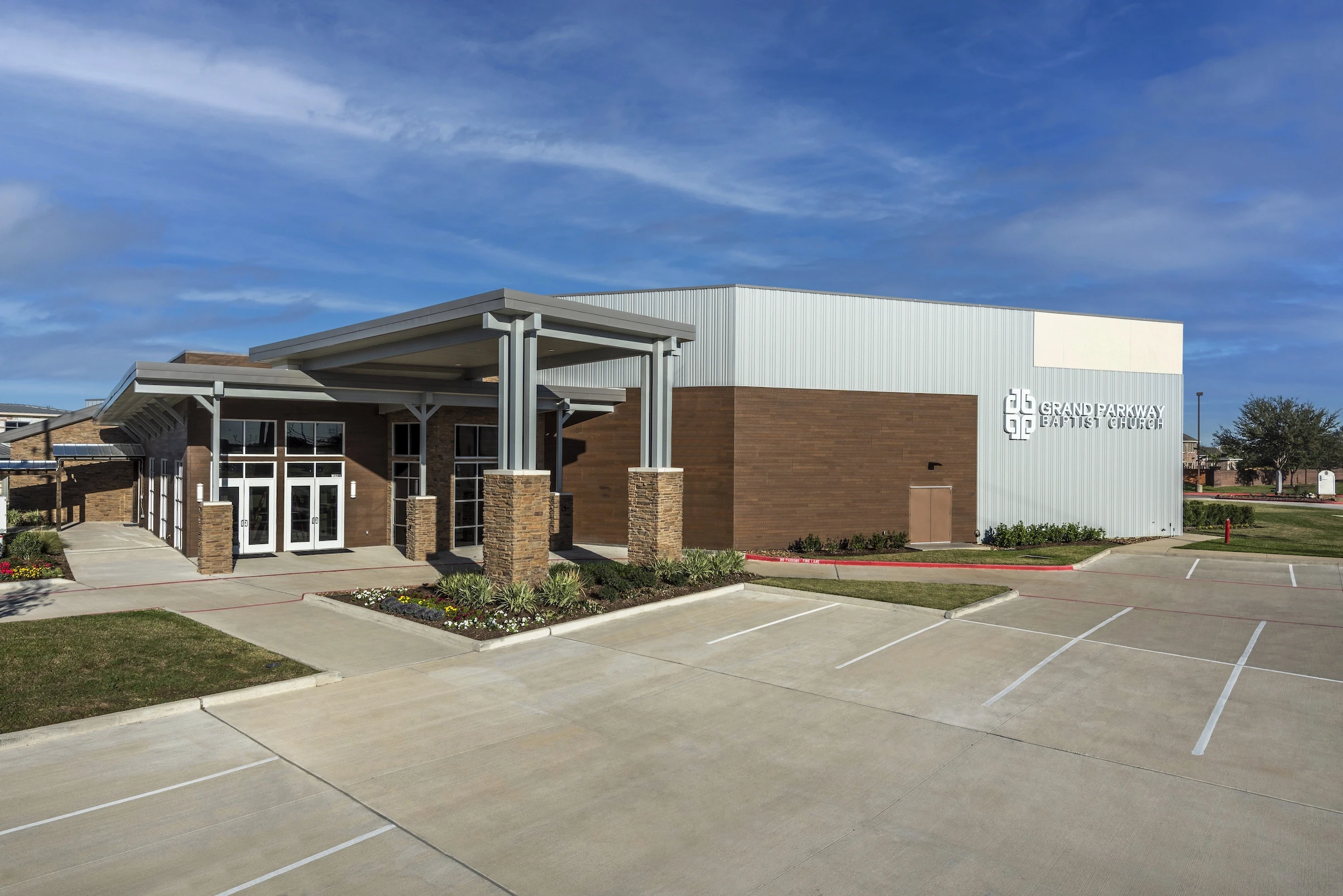 A modern, single-story building with a mix of metal and brown brick exterior sits next to an empty parking lot under a blue sky. The sign on the building reads “Grand Parkway Baptist Church,” featuring a recent addition by Meadway Construction.