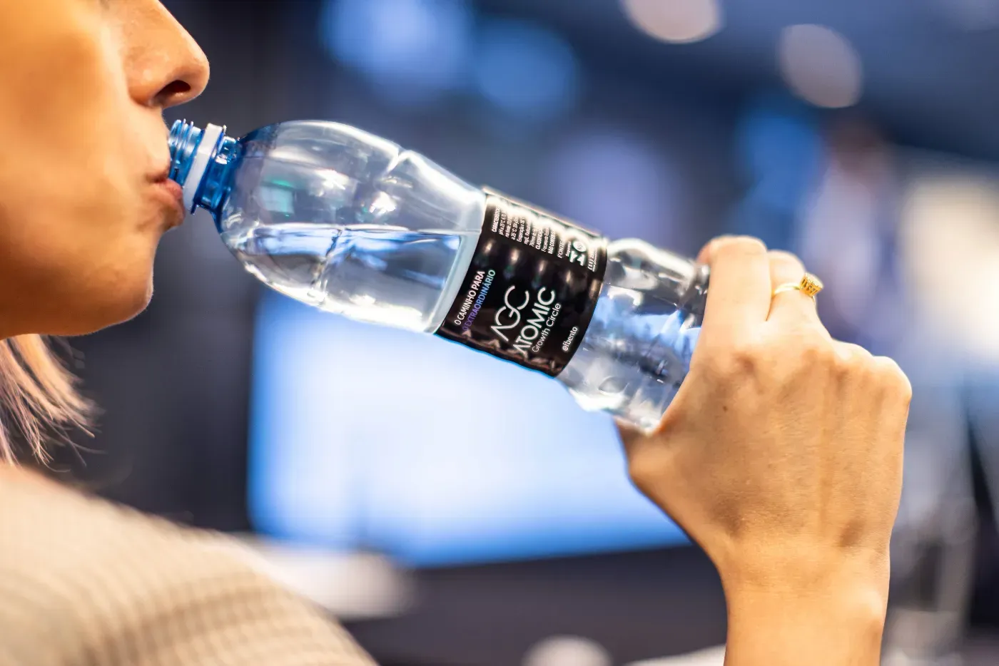A close-up of a person holding a bottle of water and taking a sip, with a blurred background. Atomic Growth Club. Atomic Group. Br24. 