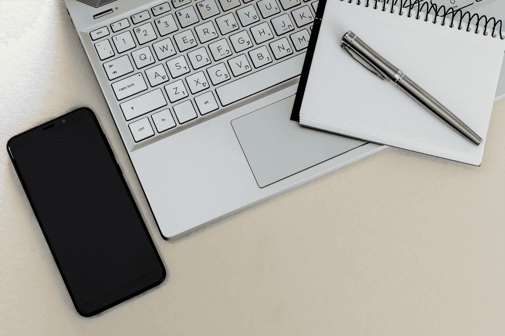 Laptop keyboard with a notebook, pen, and smartphone on a desk.