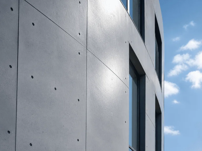 Die saubere Außenfassade eines modernen Gebäudes aus grauem Sichtbeton mit charakteristischen Schalungslöchern, die im Sonnenlicht vor einem leicht bewölkten blauen Himmel glänzt.