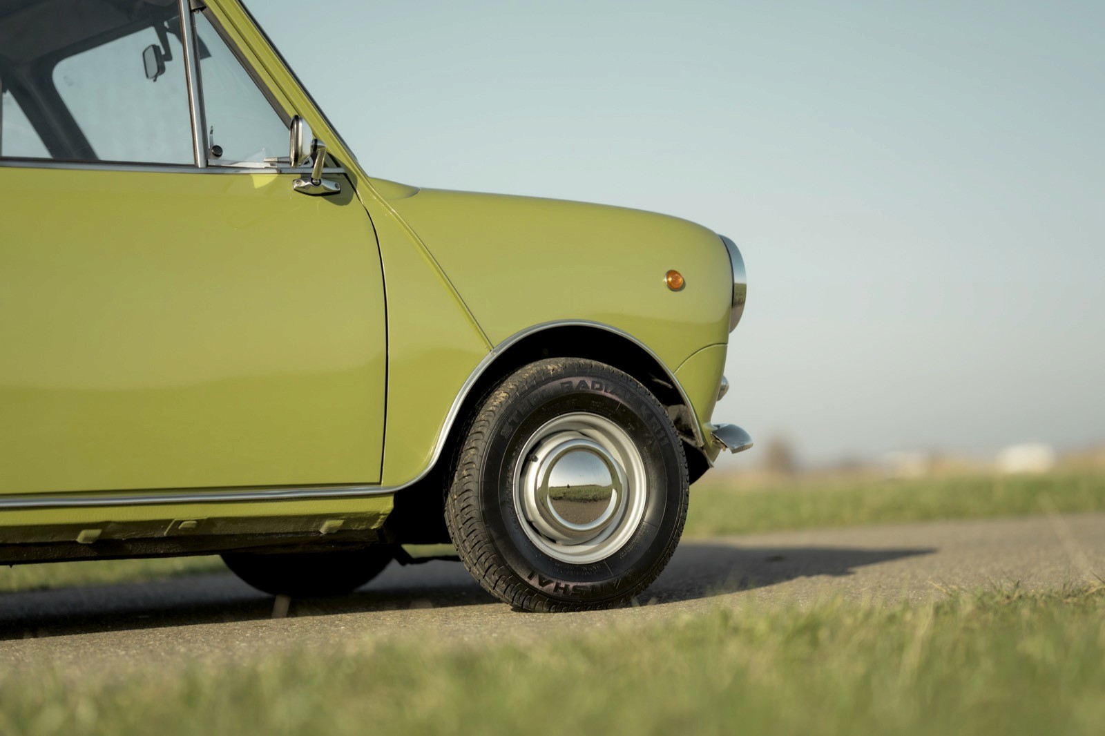 Close-up of a green vintage car parked on a gravel road with grassy fields in the background.