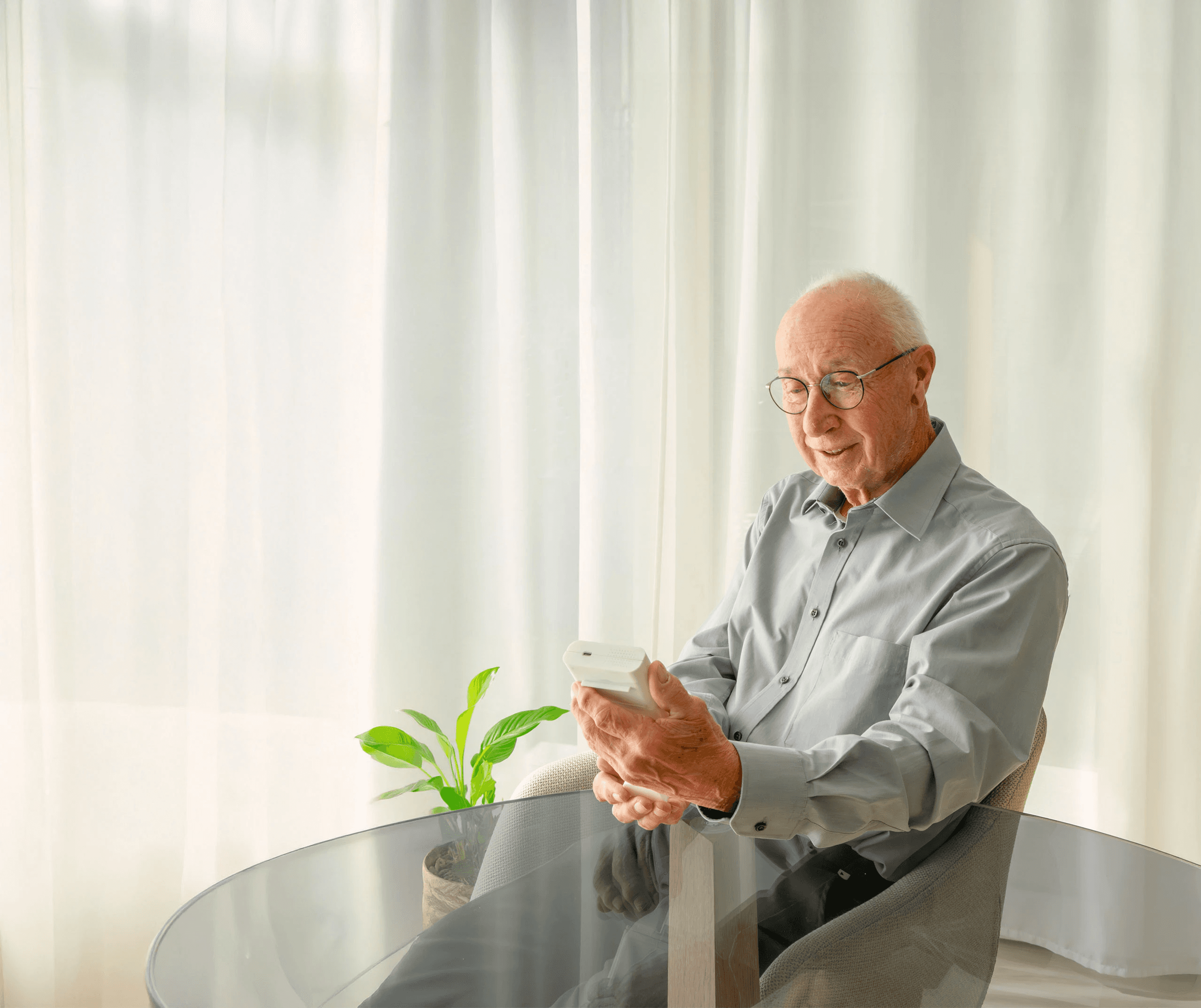 A smiling elderly man, wearing glasses, sitting at a table and holding a white smart medication dispenser (Medicase) in his hands.