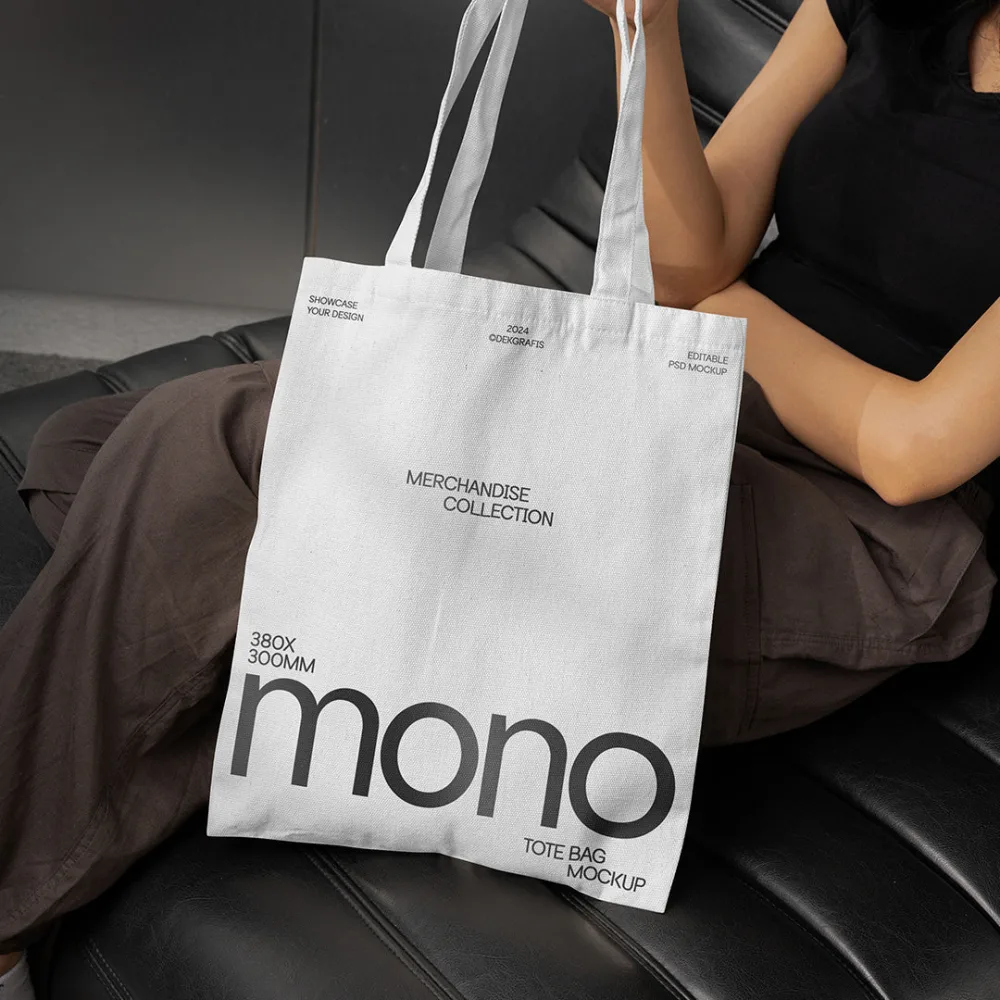 A white tote bag with a blue gradient design, featuring bold modern typography, held by a seated individual dressed in casual attire. The setting includes a sleek black leather couch in soft lighting.