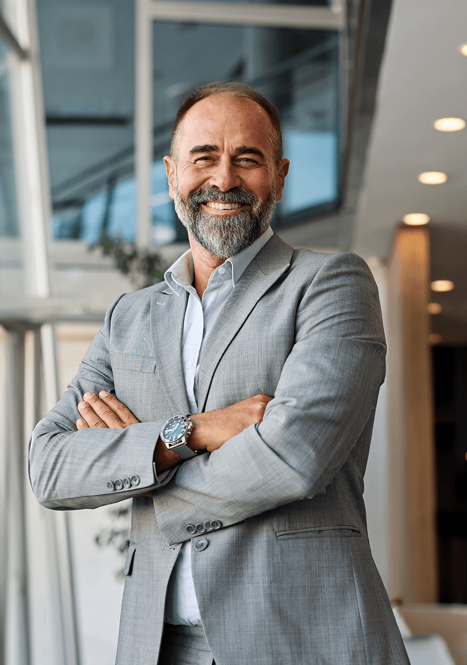 “Smiling middle-aged man with a gray beard, wearing a light gray suit, standing with arms crossed in a bright modern office.”