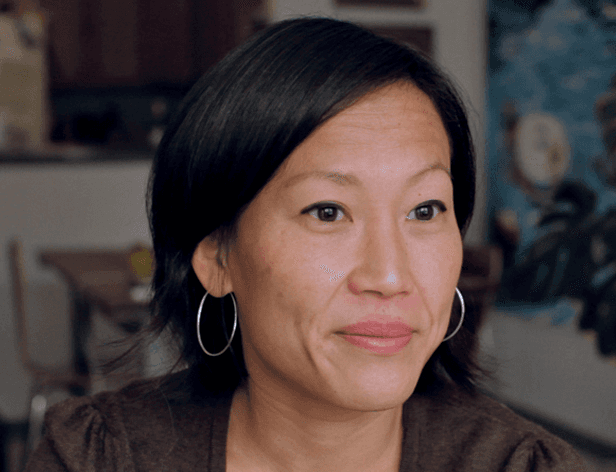 A woman with black hair and hoop earrings wears a dark brown top. She looks directly at the camera with a neutral expression. The background is out of focus, showing a portion of a wooden table and chair, and a blue artwork on the wall behind her.