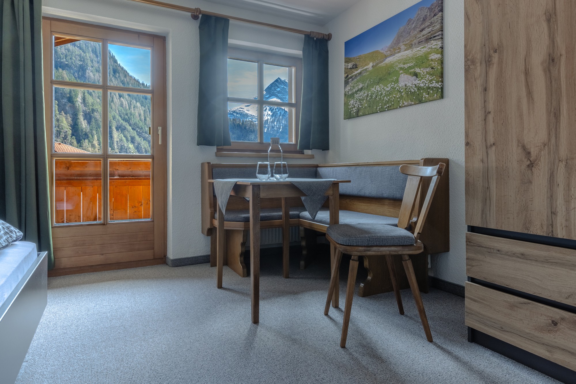 Ein gemütliches Zimmer im Alpenstil verfügt über einen kleinen Esstisch aus Holz neben einem großen Fenster mit Bergblick, ergänzt durch rustikales Dekor und natürliche Holzmöbel.
