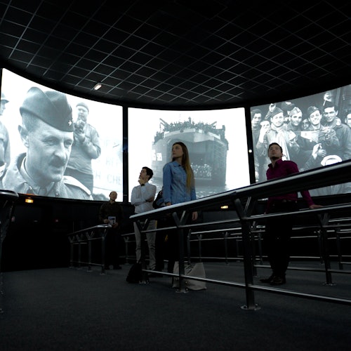 Personen, die sich historische Schwarz-Weiß-Kriegsaufnahmen auf großen, gebogenen Bildschirmen in einem modernen Ausstellungsraum mit dunkler Decke ansehen.