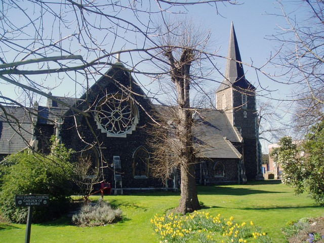 A smiling man with curly hair is sitting at a desk, looking at the camera, with a laptop and a lamp in the background.