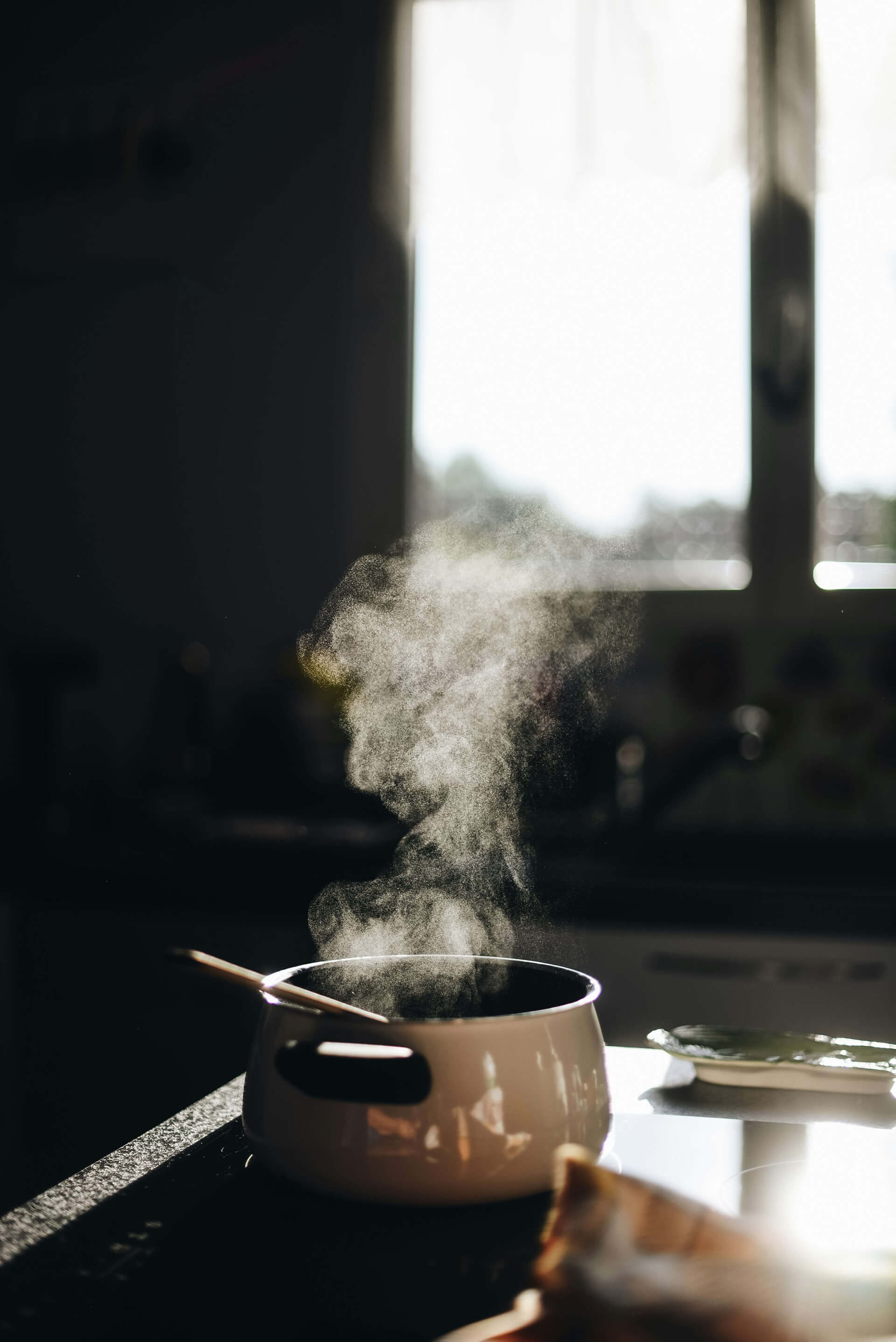 Steam rises from a white pot on a dark kitchen counter, softly backlit by sunlight streaming through a window. A wooden spoon rests inside the pot, suggesting something freshly cooked or simmering in a cozy home setting.