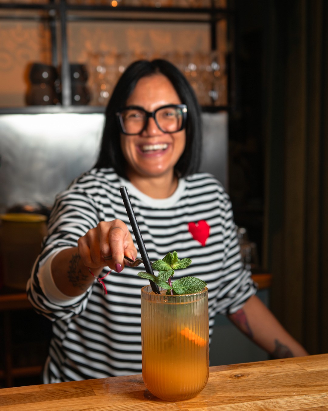Eine lächelnde Frau mit dunklem Haar und Brille hält ein Minz-Limetten-Getränk mit einem Strohhalm, sie trägt ein gestreiftes Hemd mit einem roten Herzen.