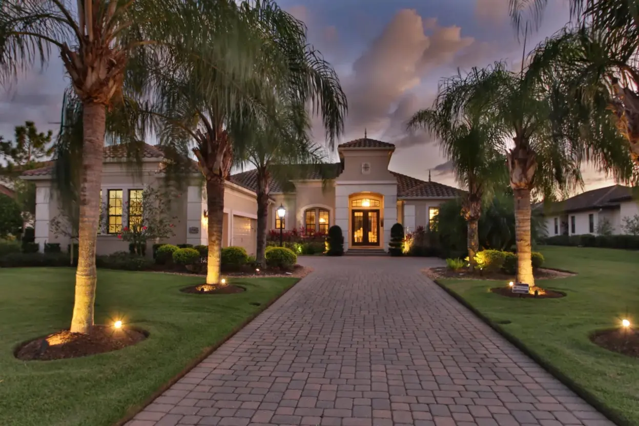 A luxurious house in Bradenton, FL, features a beautifully landscaped front yard with tall palm trees lining a paved driveway leading to the entrance, illuminated by soft, ambient lighting at dusk.