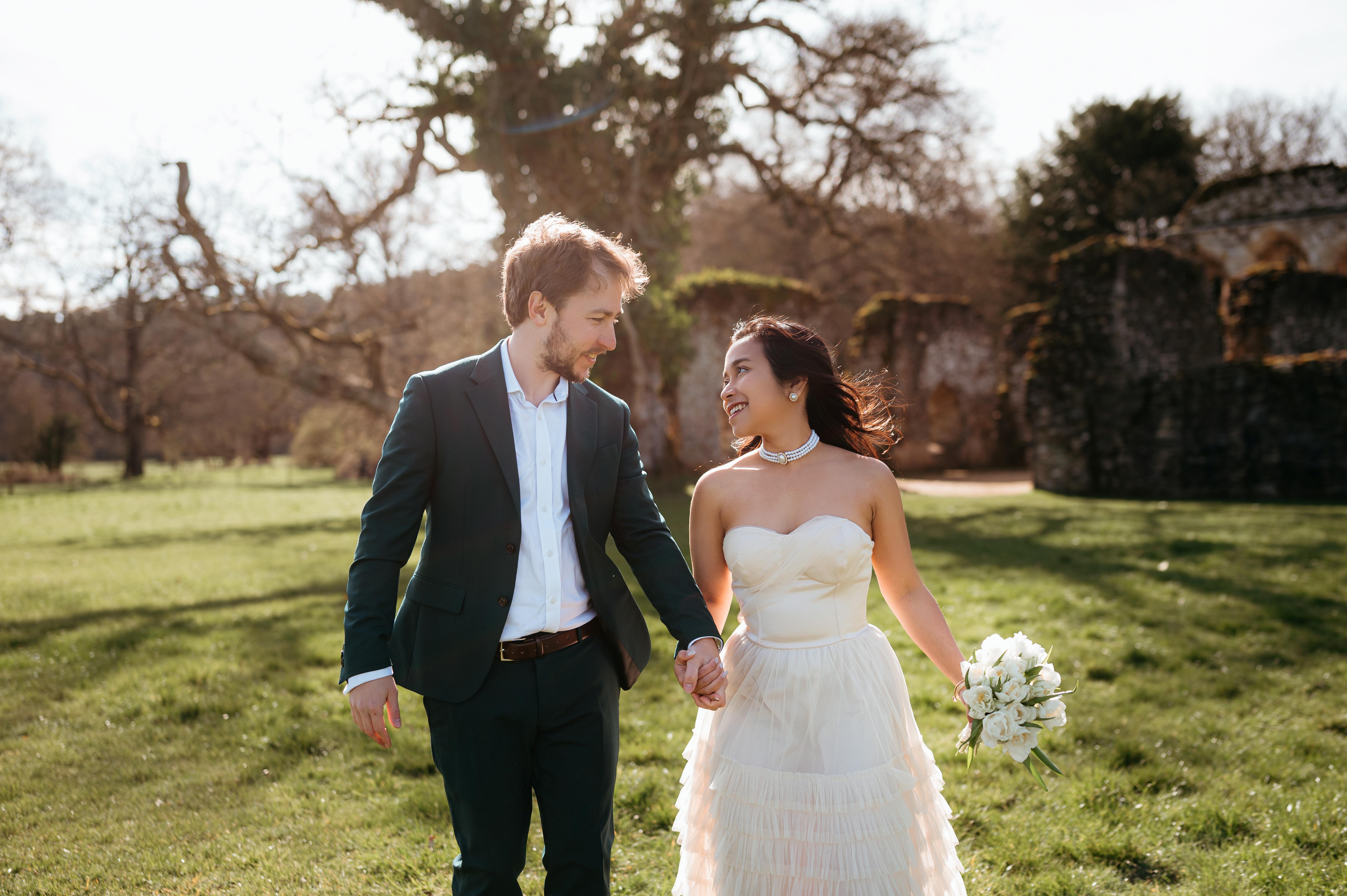 A bride and groom hold hands tenderly, the bride's bouquet of dried flowers hanging low. They stand in a softly lit, elegant interior, conveying romance.