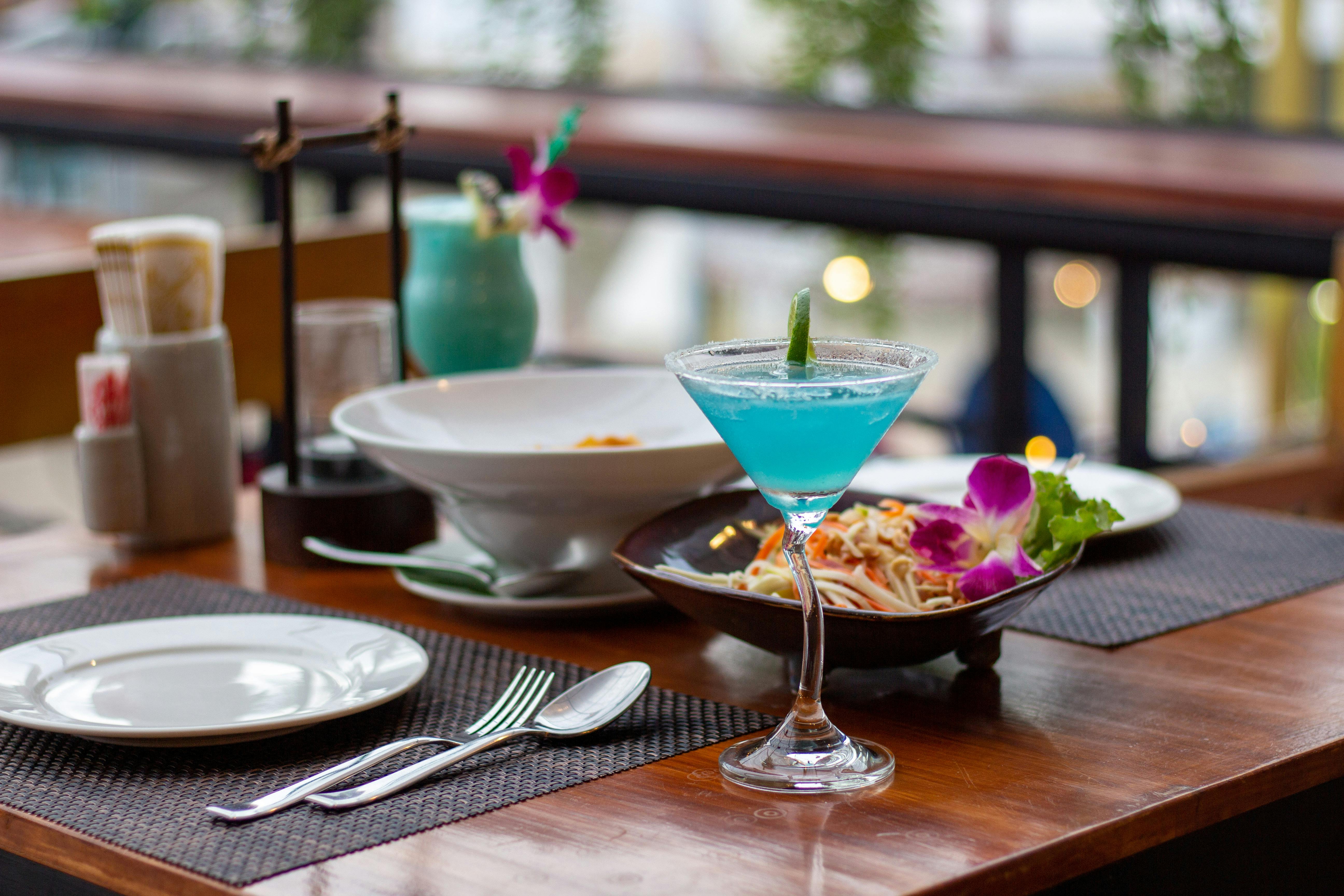 Outdoor restaurant table with blue cocktail, plated meal, and table setting in a casual dining