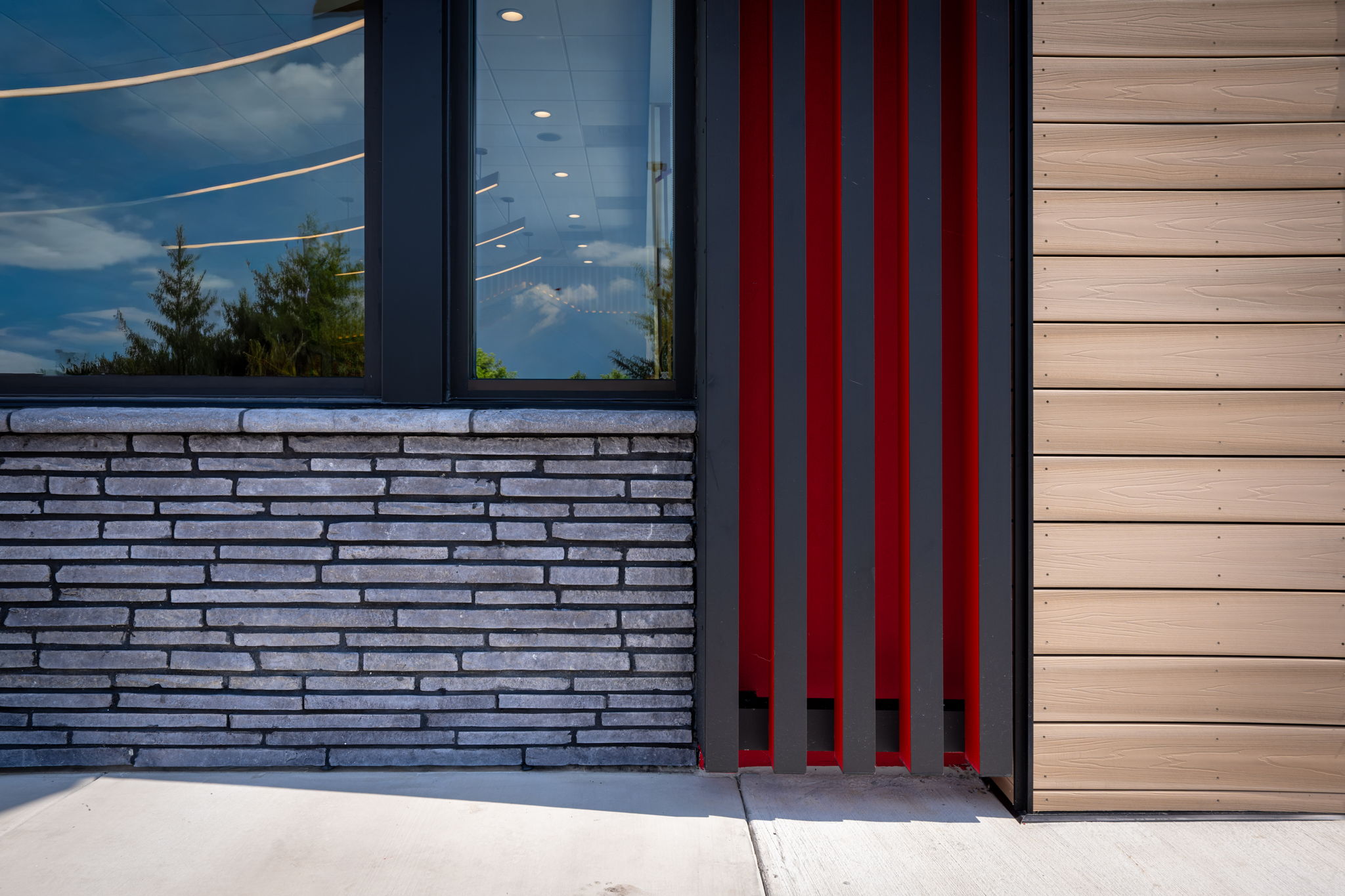 A modern commercial building exterior featuring a grey linear stone veneer wall, vertical red architectural accents, and light wood-textured siding next to a large black-framed window.