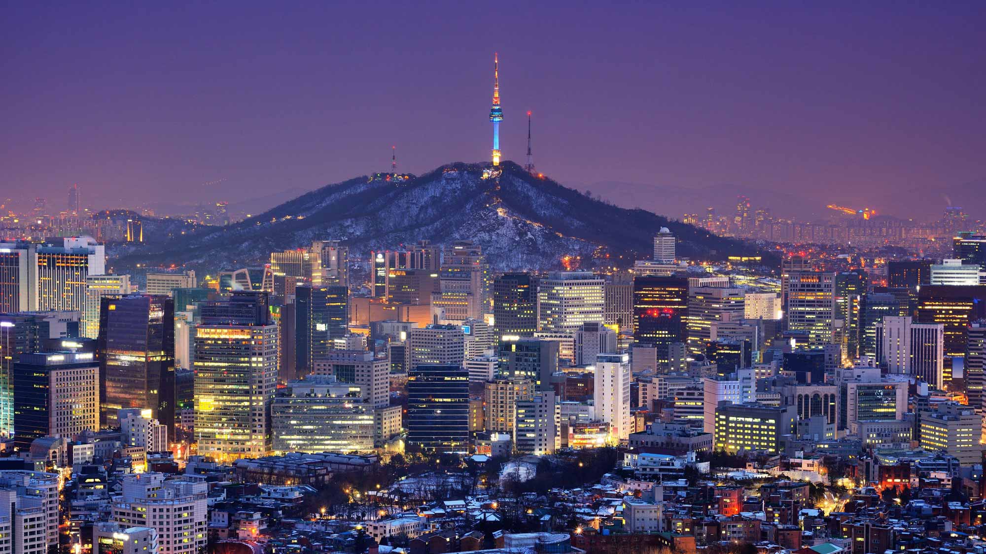 City skyline at night with illuminated buildings and a hilltop tower in the background.