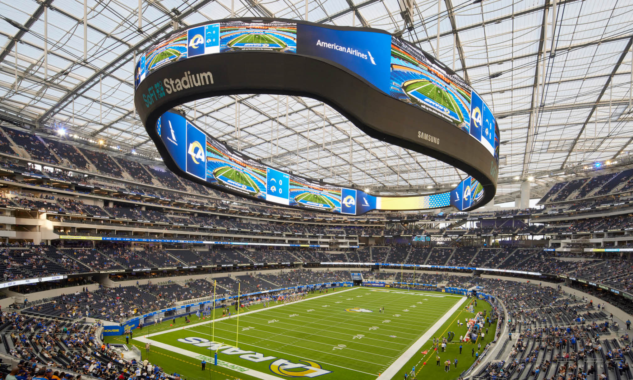Wide view of SoFi Stadium during an NFL game showing the field, crowd, and massive circular video board overhead.