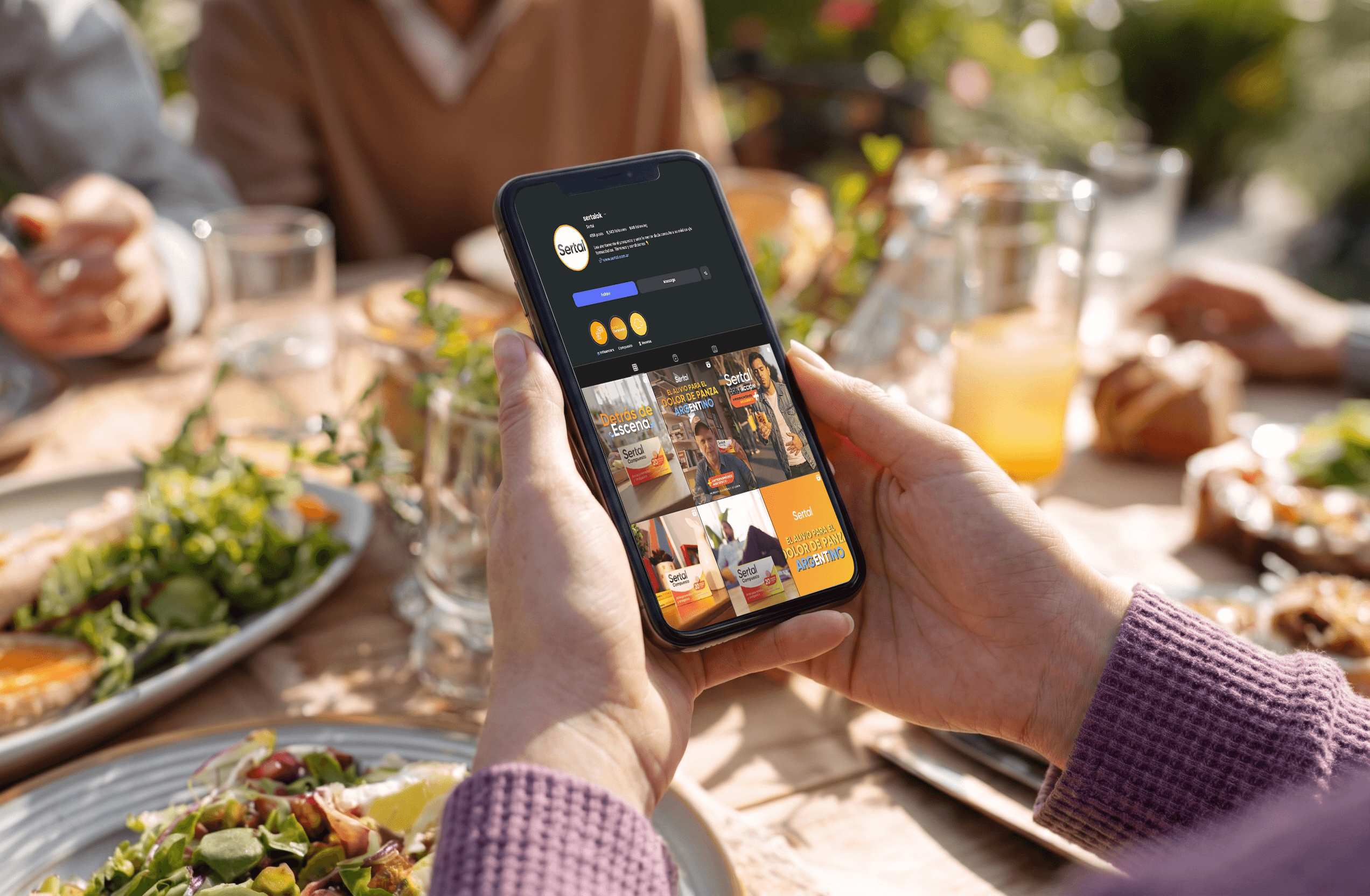 A person holding a smartphone displaying the feed of the brand Sertal over a outdoor table with vibrant and delicious food.