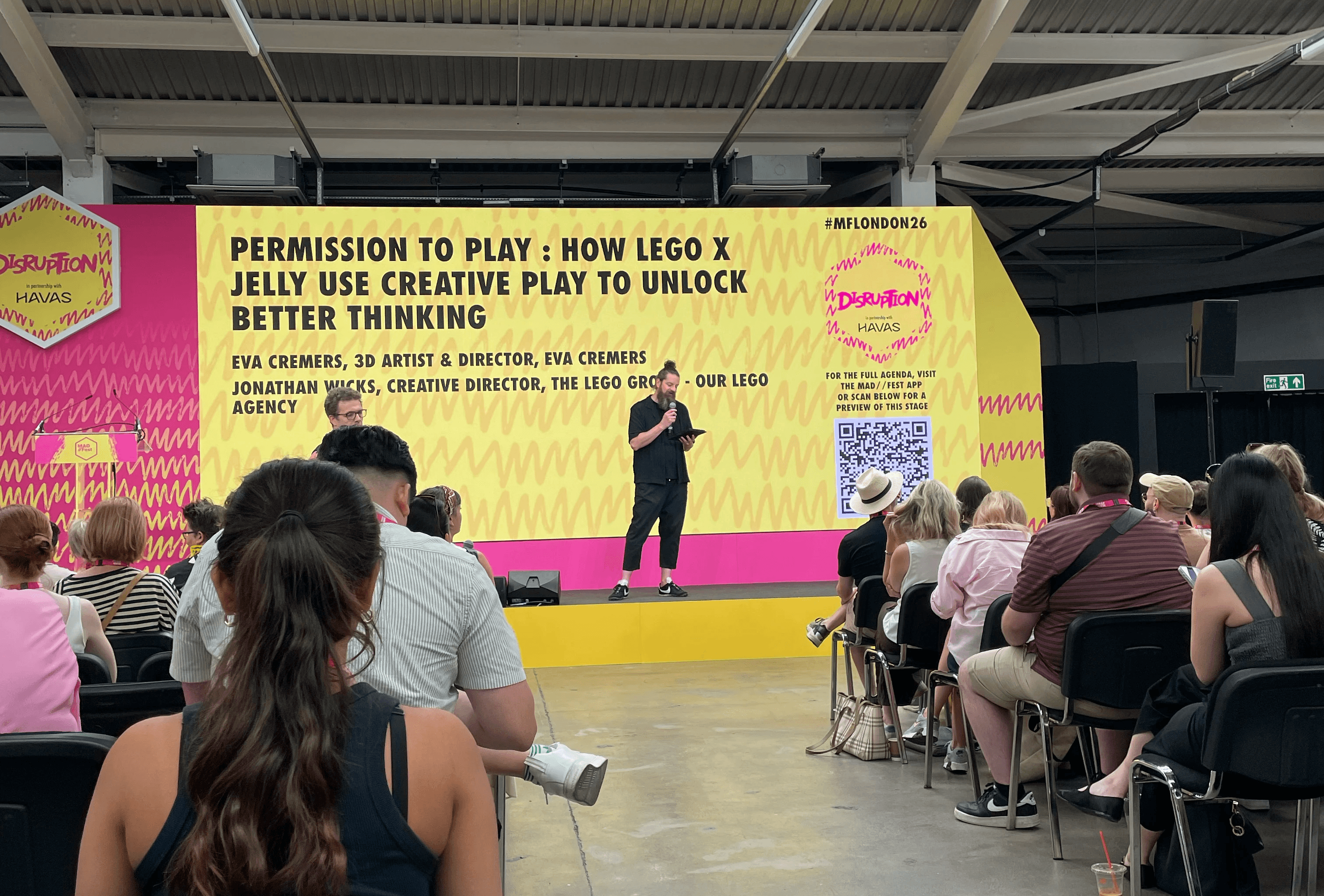 A speaker addresses a seated audience at MADFEST 2026's Disruption stage, in front of a yellow and pink screen titled 'Permission to Play: How Lego x Jelly Use Creative Play to Unlock Better Thinking.'