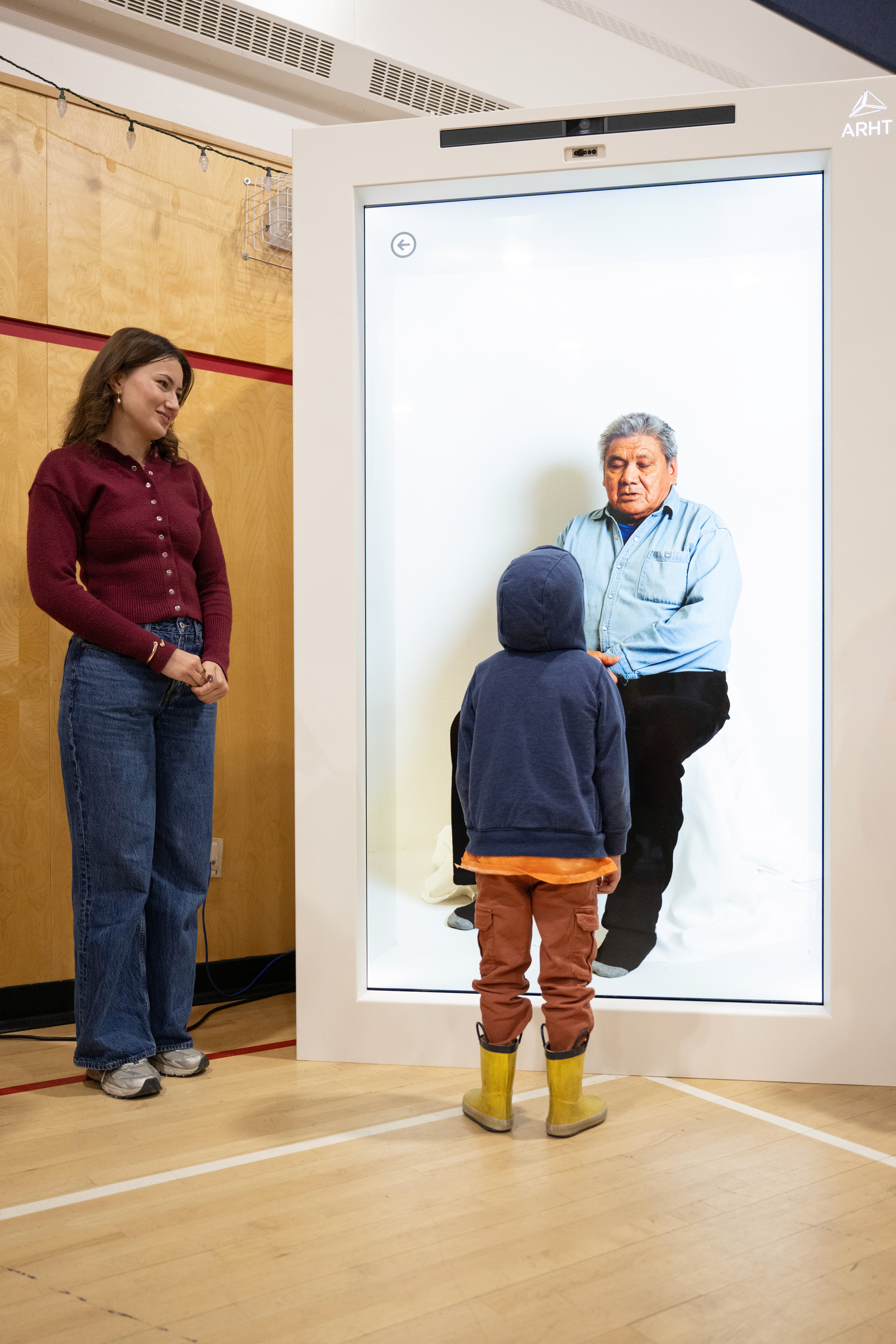 A young child in a blue hoodie stands face to face with a life-size holographic projection of an Elder man seated and looking forward. The intimate scale of the interaction suggests quiet conversation across generations.