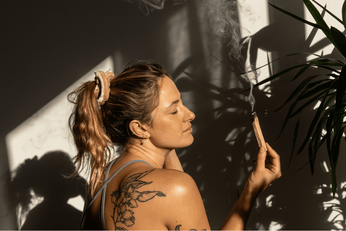 A woman with tattoos holds a smoldering incense stick, creating a mindful atmosphere in a sunlit room filled with leafy plants.