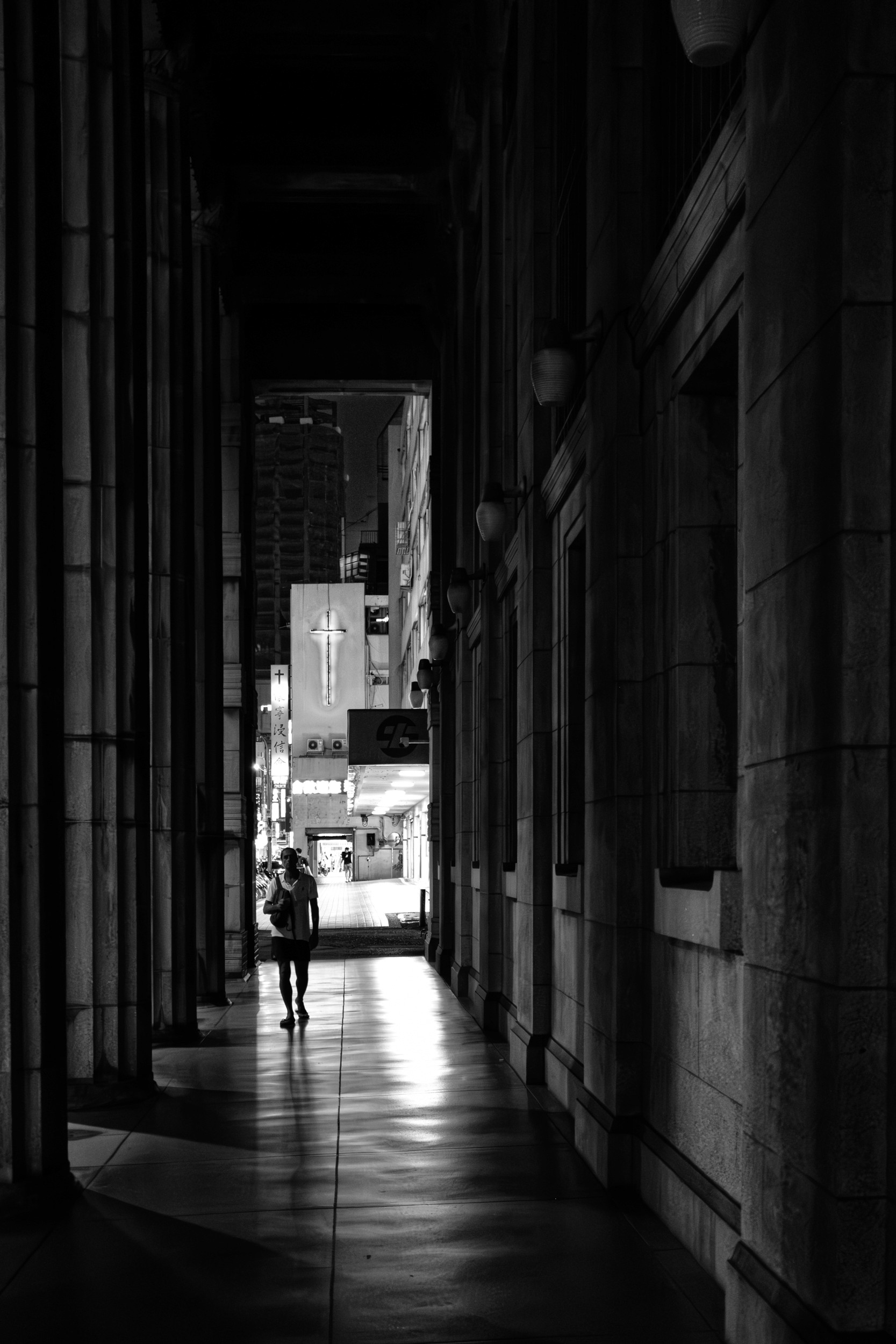 Black and white photograph of a person walking down a long dark colonnade at night toward a brightly lit street.