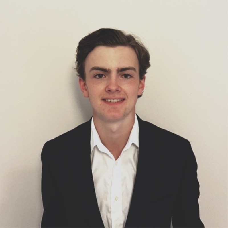A focused woman with long red hair is seated at a desk, wearing a black blazer and dark blouse. She intently looks at camera, smiling slightly, with an open laptop in front of her. The setting is a warm and serene office setup in the background. Soft daylight filters through, creating a calm atmosphere. The composition captures her in a frontal profile with a clear emphasis on her concentration and the serene indoor setting.