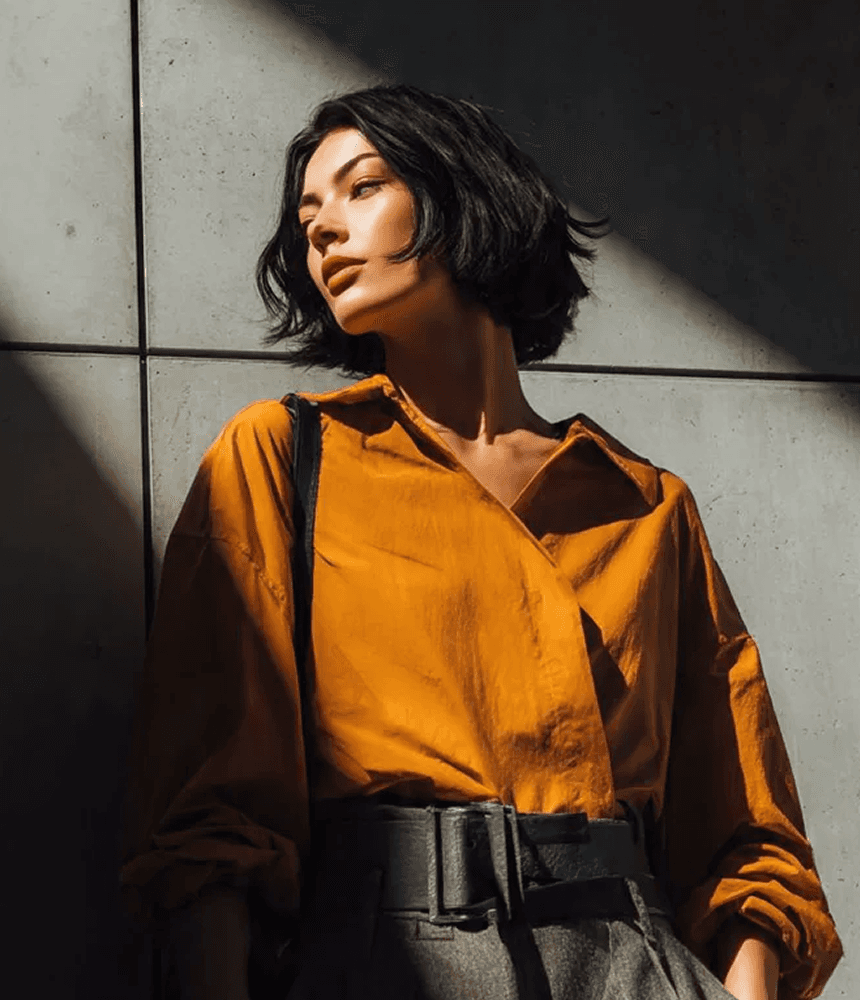 A woman in a yellow shirt and gray pants leans against a wall, appearing relaxed and casual.