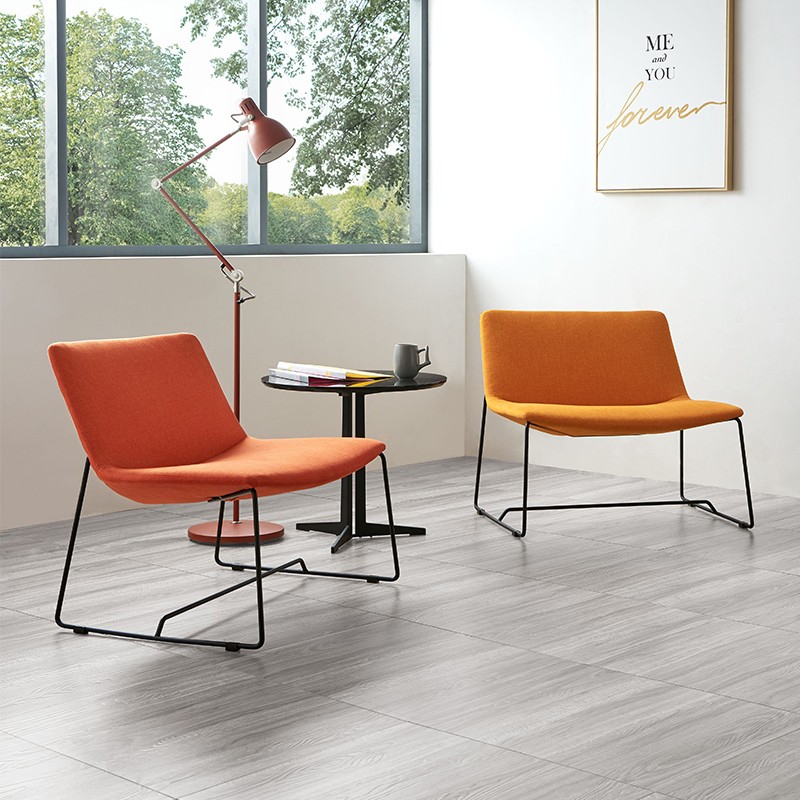 Modern minimalist lounge area featuring two low-profile upholstered chairs in orange and yellow, a slim black side table, and a red industrial floor lamp against a bright window and white wall with framed art.