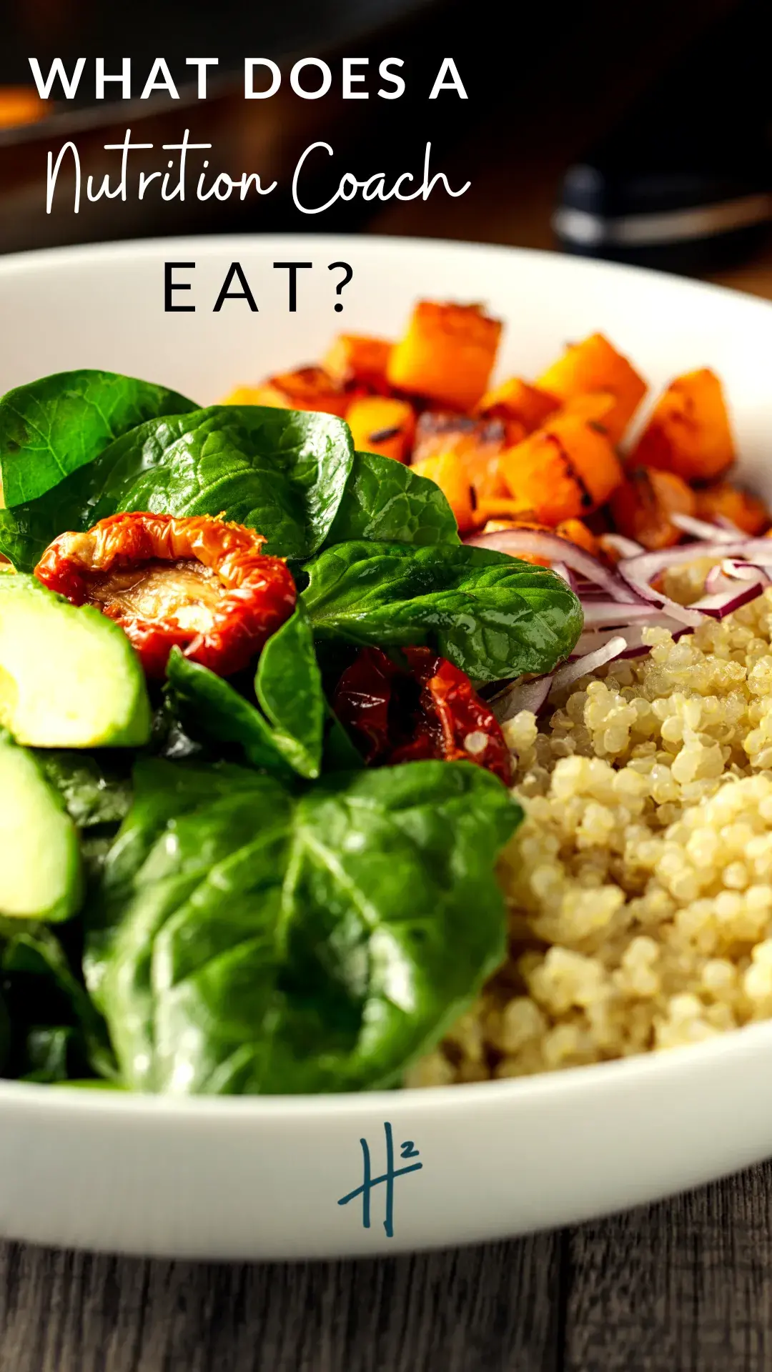 A bowl filled with quinoa, fresh spinach, sliced cucumbers, roasted sweet potatoes, red onions, and sun-dried tomatoes, set on a wooden table with text overlay reading "What Does a Nutrition Coach Eat?".