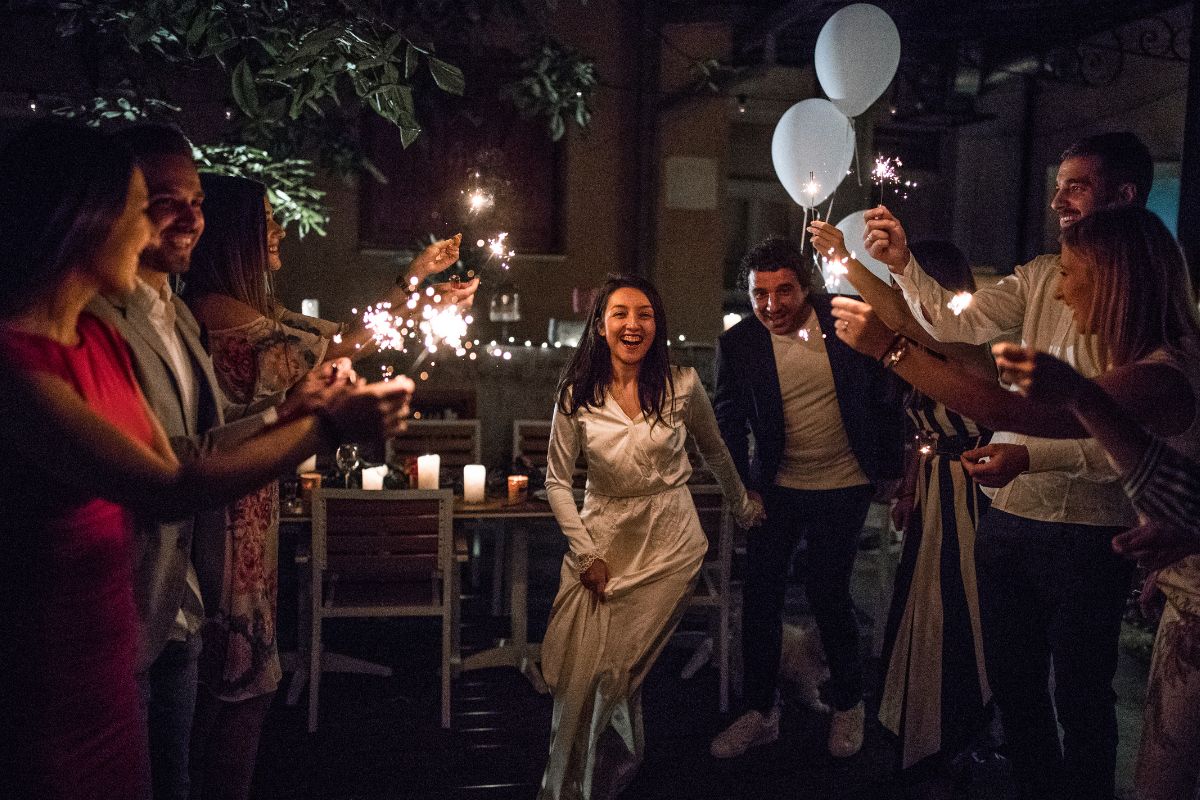 A magical outdoor wedding reception at night, where the bride and groom walk hand in hand through a tunnel of glowing sparklers held by cheering guests