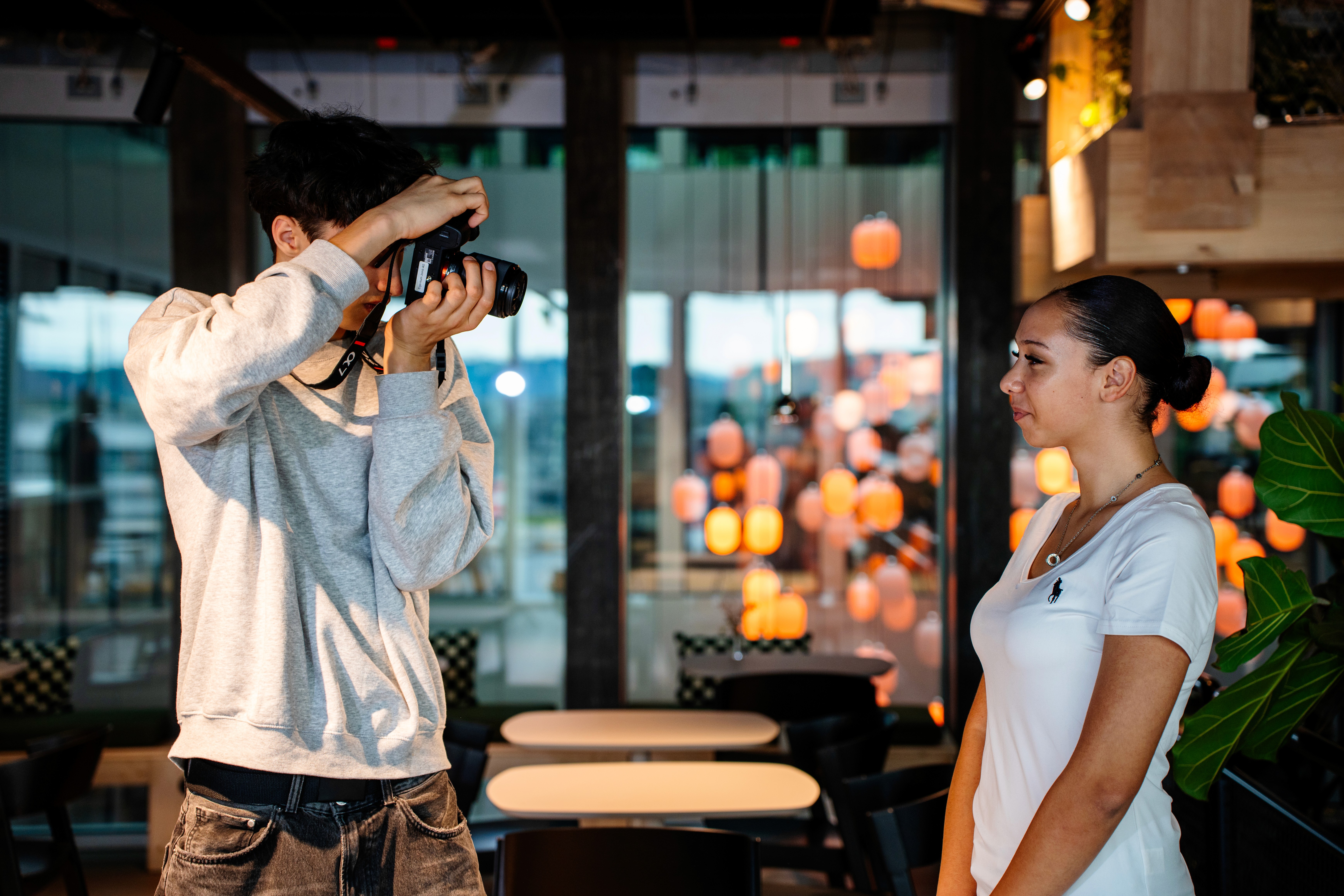A young person with curly hair smiles at the camera while sitting in front of a laptop against a dark background.