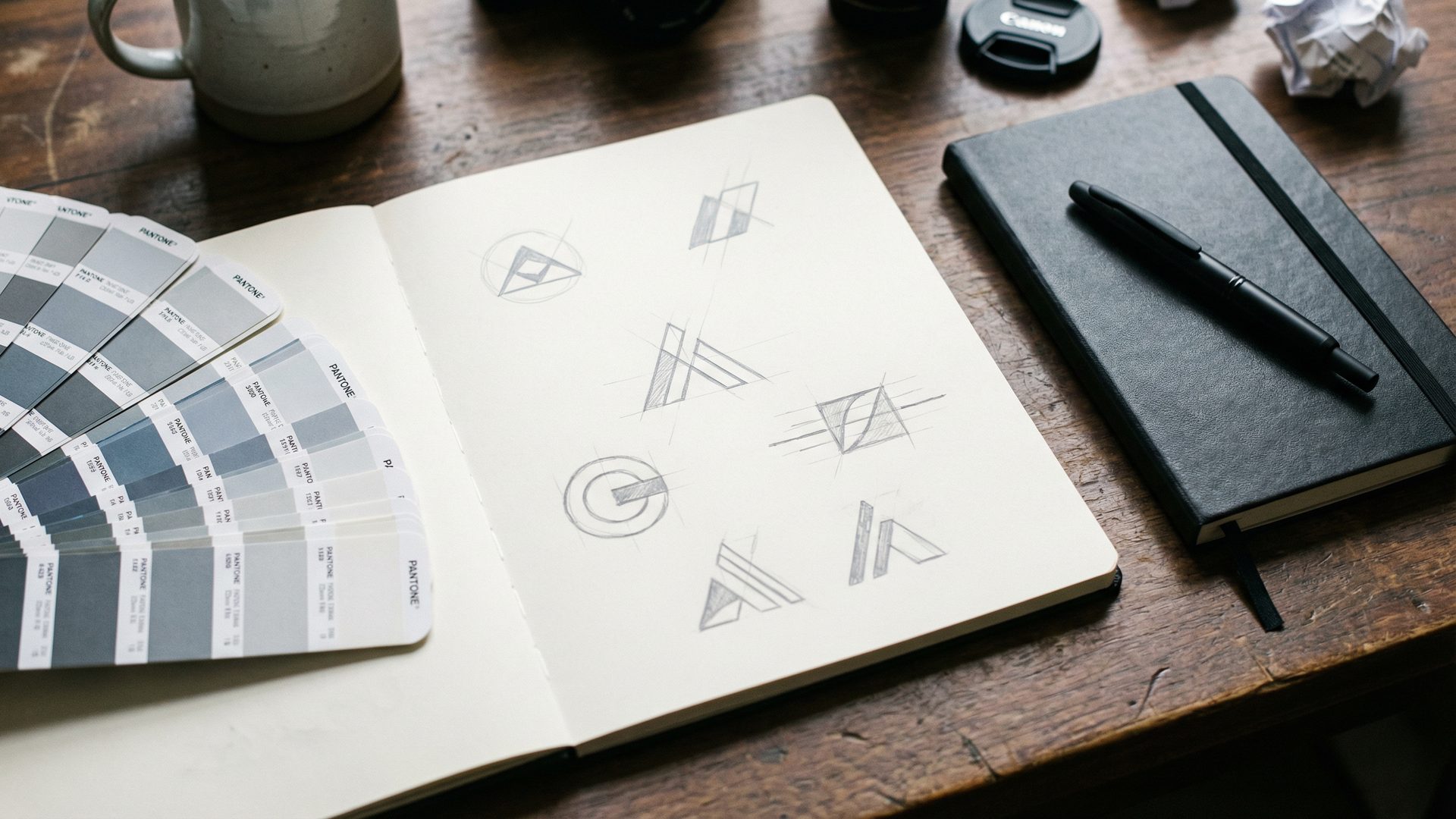 A sketchbook open to a page with pencil drawings of minimalist tech company logo ideas, surrounded by a fanned Pantone swatch book in cool greys and slate blues, and a closed black Moleskine notebook with a pen on a dark walnut desk.